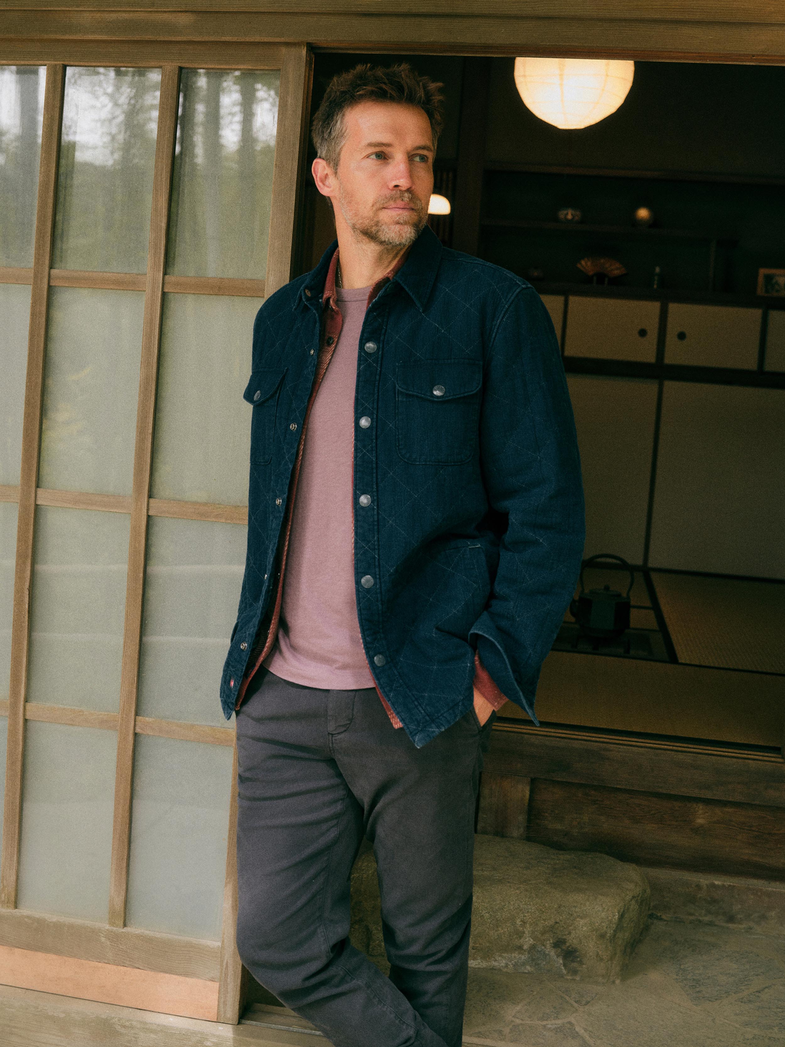 A man wearing a dark indigo denim overshirt over a dusty pink t-shirt and dark gray trousers, standing in a doorway of a traditional Japanese-style house.