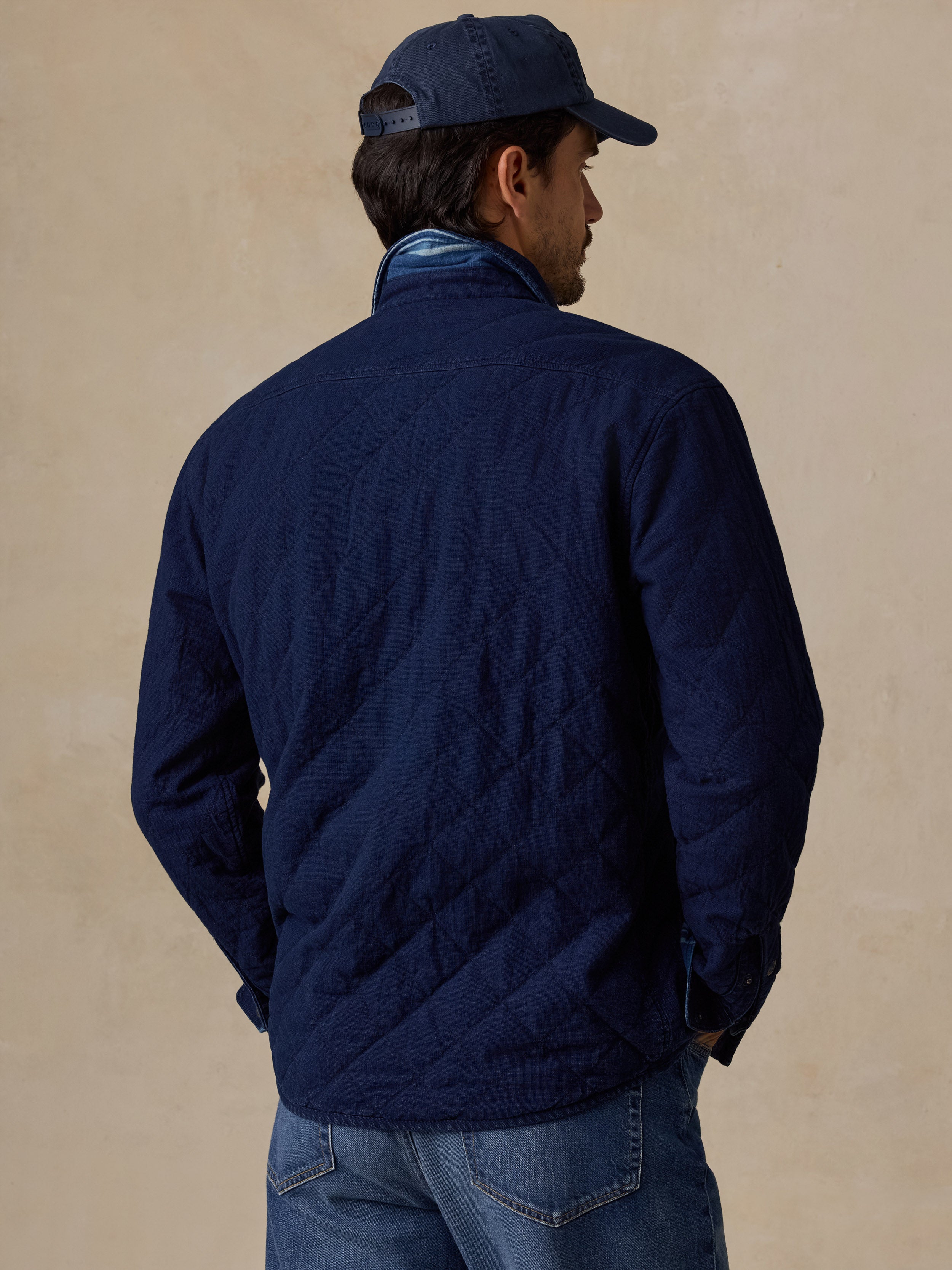 A man wearing a navy blue quilted jacket, blue jeans, and a navy blue baseball cap, shown from the back against a textured beige wall.