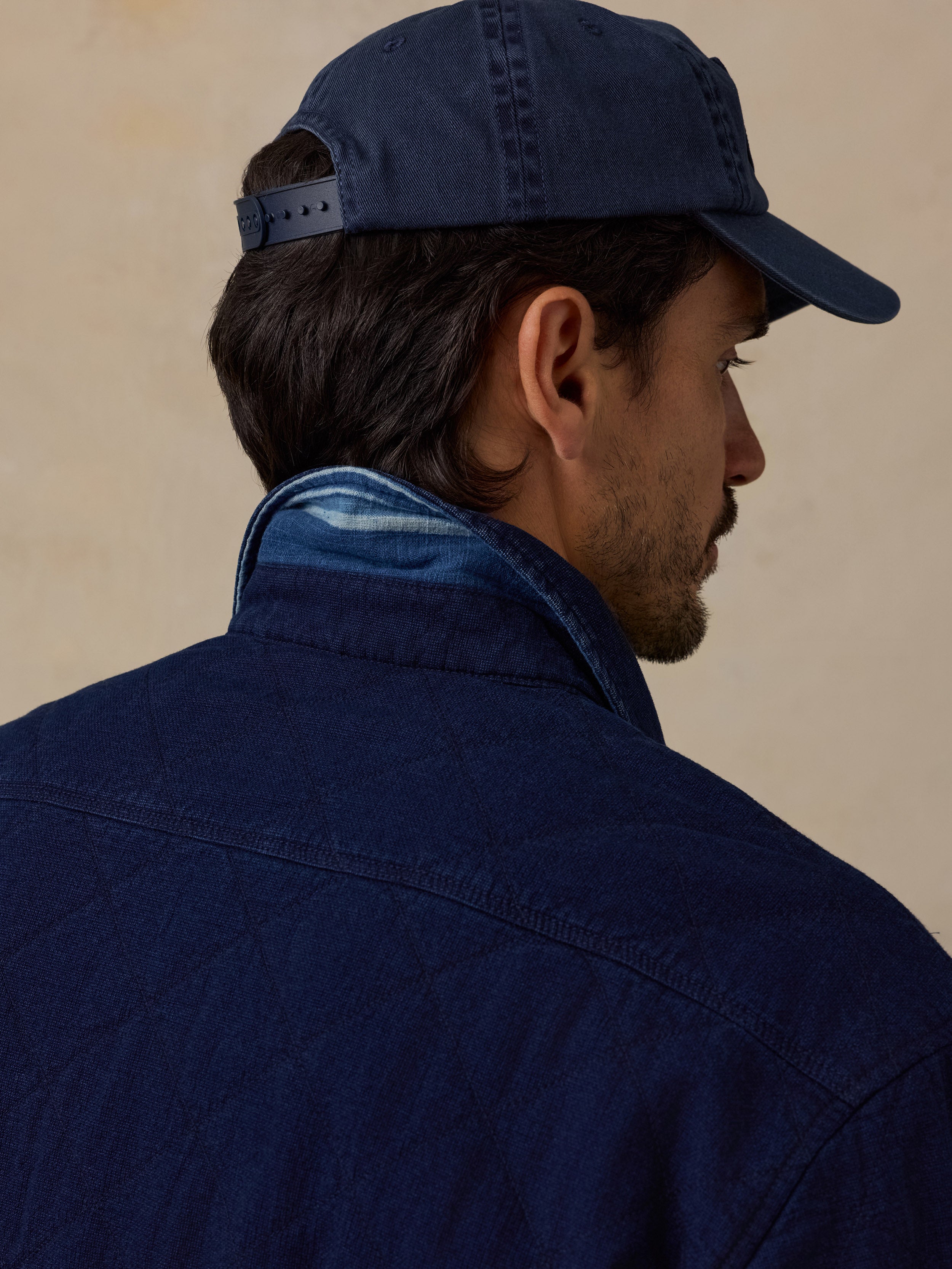 A man shown from the back and side profile wearing a dark indigo quilted overshirt, a matching dark indigo baseball cap, and a blue striped knit collar under his shirt collar, set against a warm beige background.