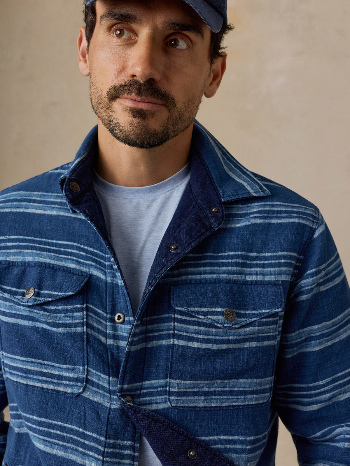 A man wearing a blue and white striped button-up shirt over a light blue t-shirt, paired with a dark blue baseball cap, set against a neutral beige background.