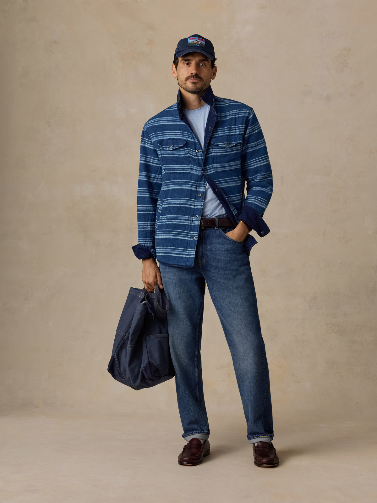 A man stands against a textured beige studio backdrop wearing a blue striped button-up shirt over a light blue T-shirt, blue jeans, brown shoes, and a navy cap, while holding a navy tote bag.