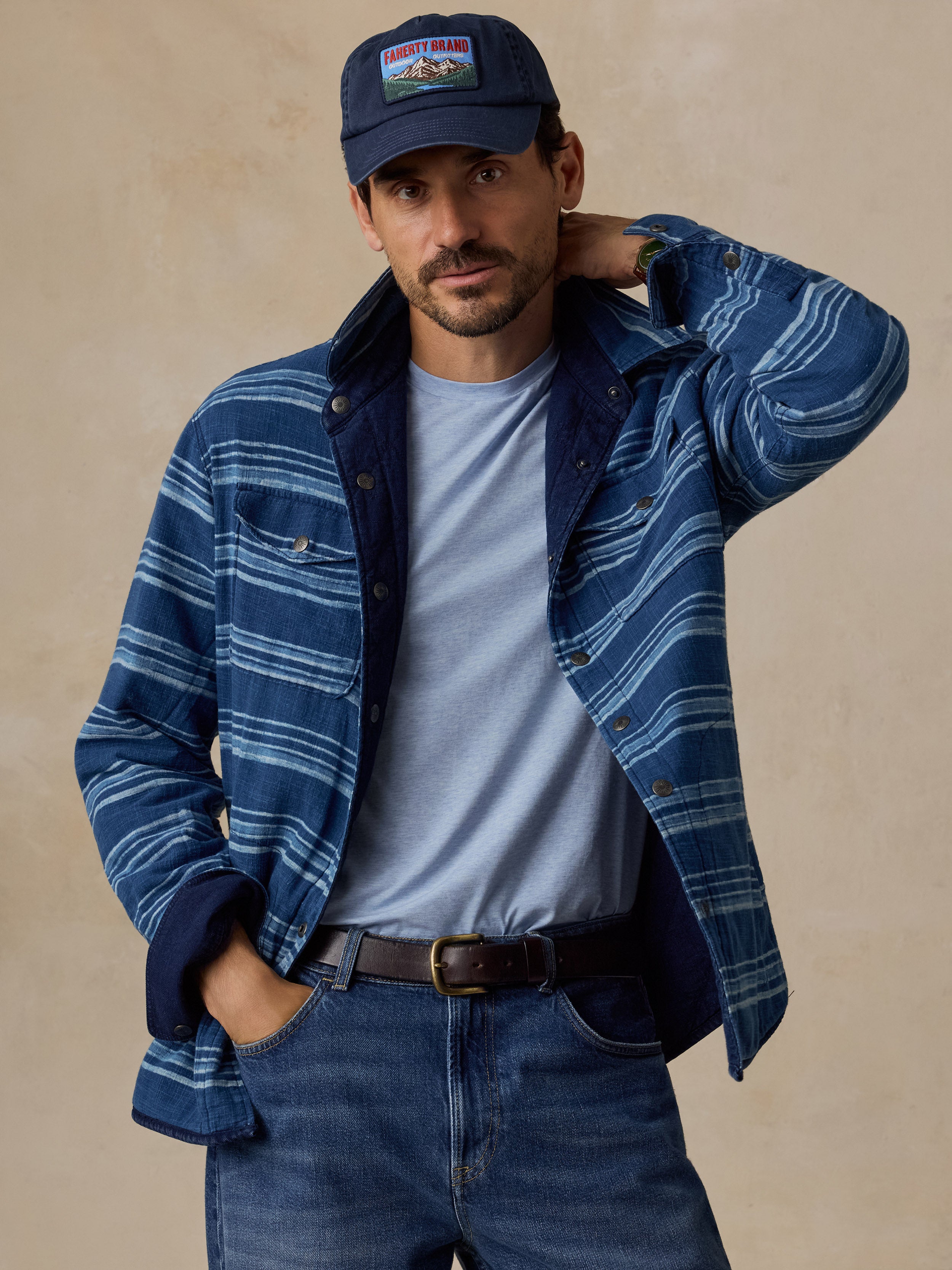 A man wearing a blue and navy striped button-up overshirt over a light blue t-shirt, paired with blue jeans and a navy baseball cap with a 'Mountain' patch, posed against a textured beige background.