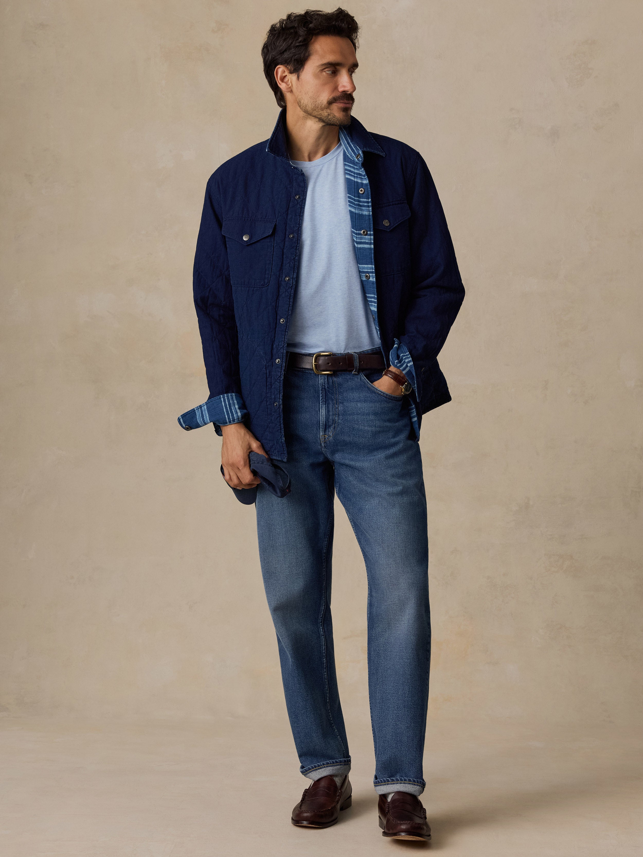 A man stands against a textured beige studio backdrop wearing a dark navy overshirt over a light blue T-shirt, blue jeans, a brown belt, and dark brown shoes.