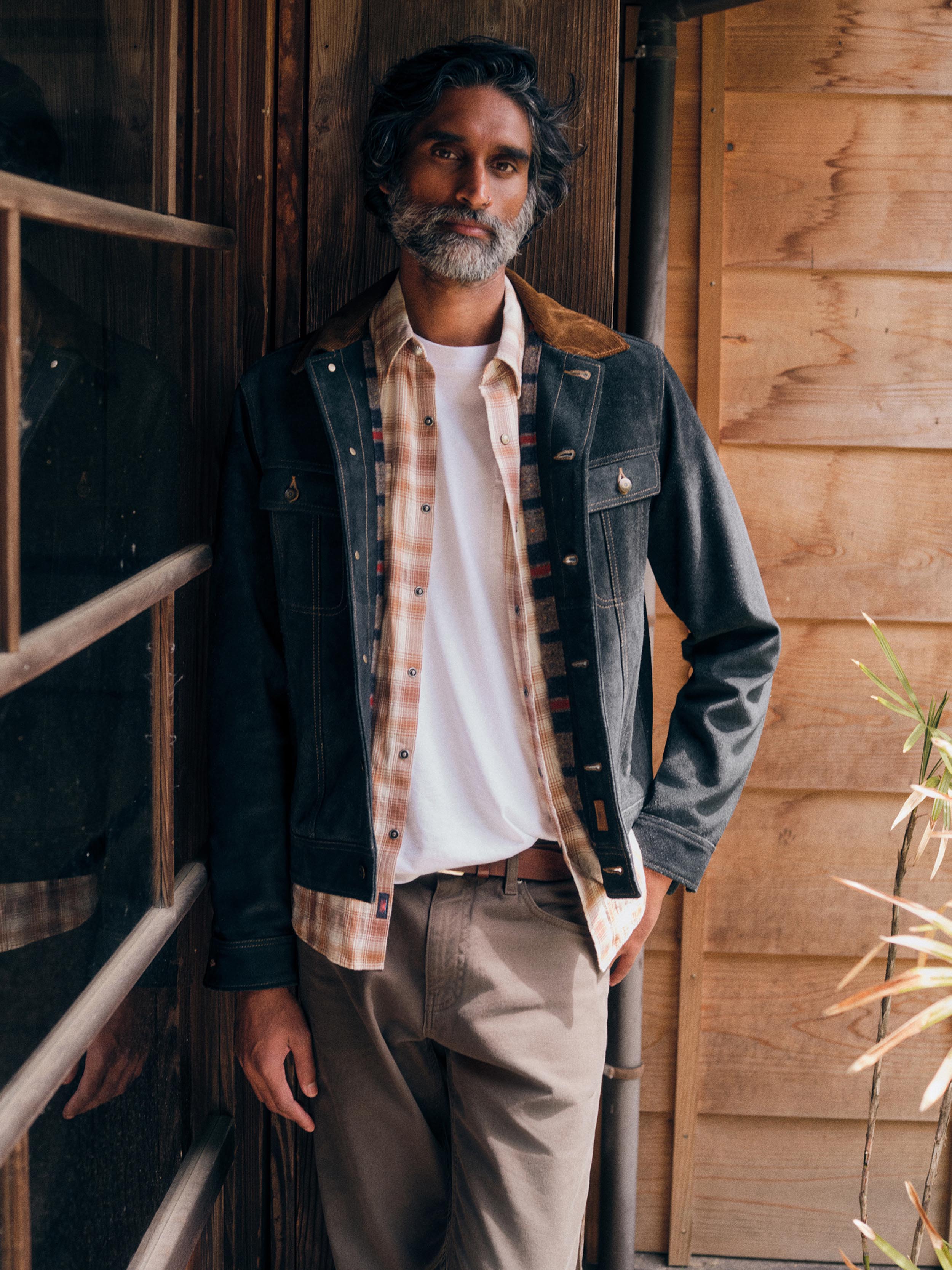 A man with a graying beard and dark hair stands in front of a rustic wooden door, wearing a dark charcoal suede jacket with brown collar lining over a beige and orange plaid flannel shirt, a white t-shirt, and khaki pants.
