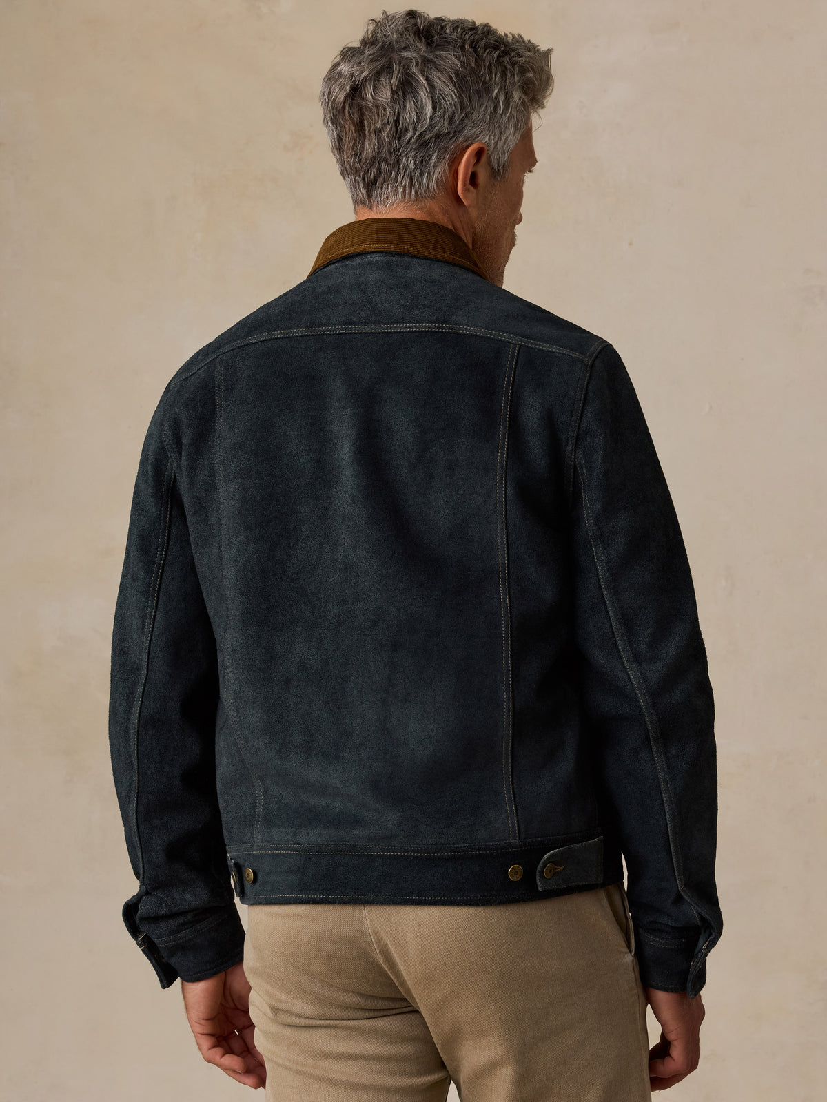 A man viewed from behind wearing a dark navy blue suede jacket with a brown corduroy collar and beige trousers, set against a textured beige background.