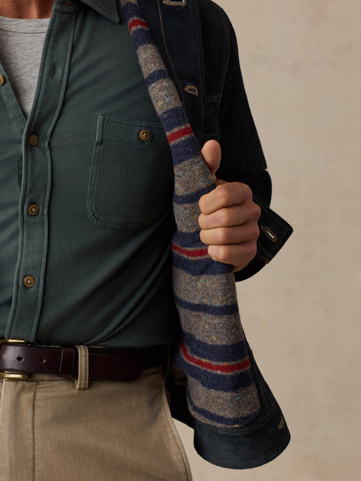 A close-up of a man wearing a dark green button-up shirt, beige trousers, and a dark overshirt, holding a striped wool sweater draped over their shoulder against a neutral beige background.