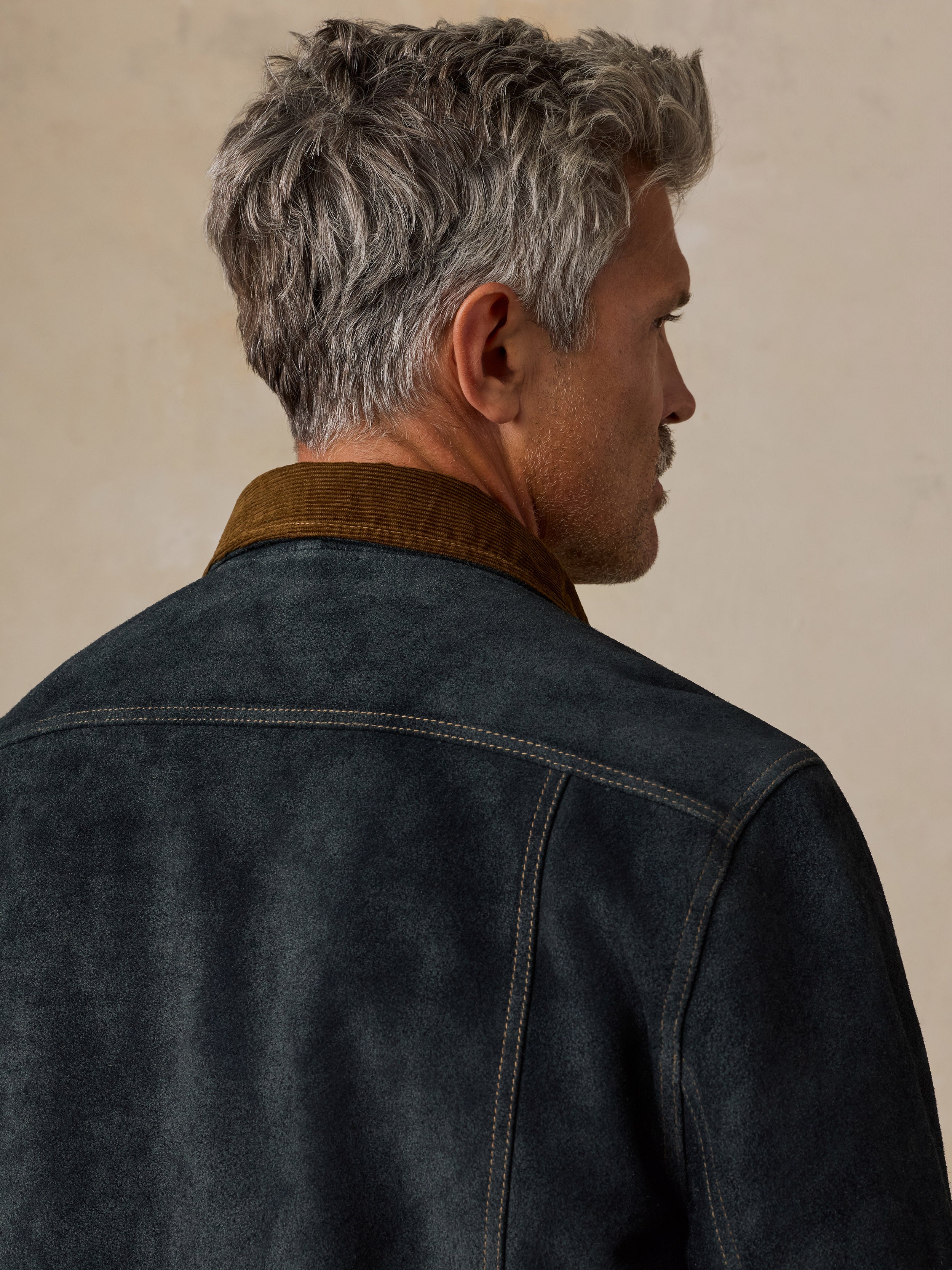 A man shown from the back and side profile wearing a dark charcoal gray suede jacket with a brown corduroy collar, set against a warm beige textured background.