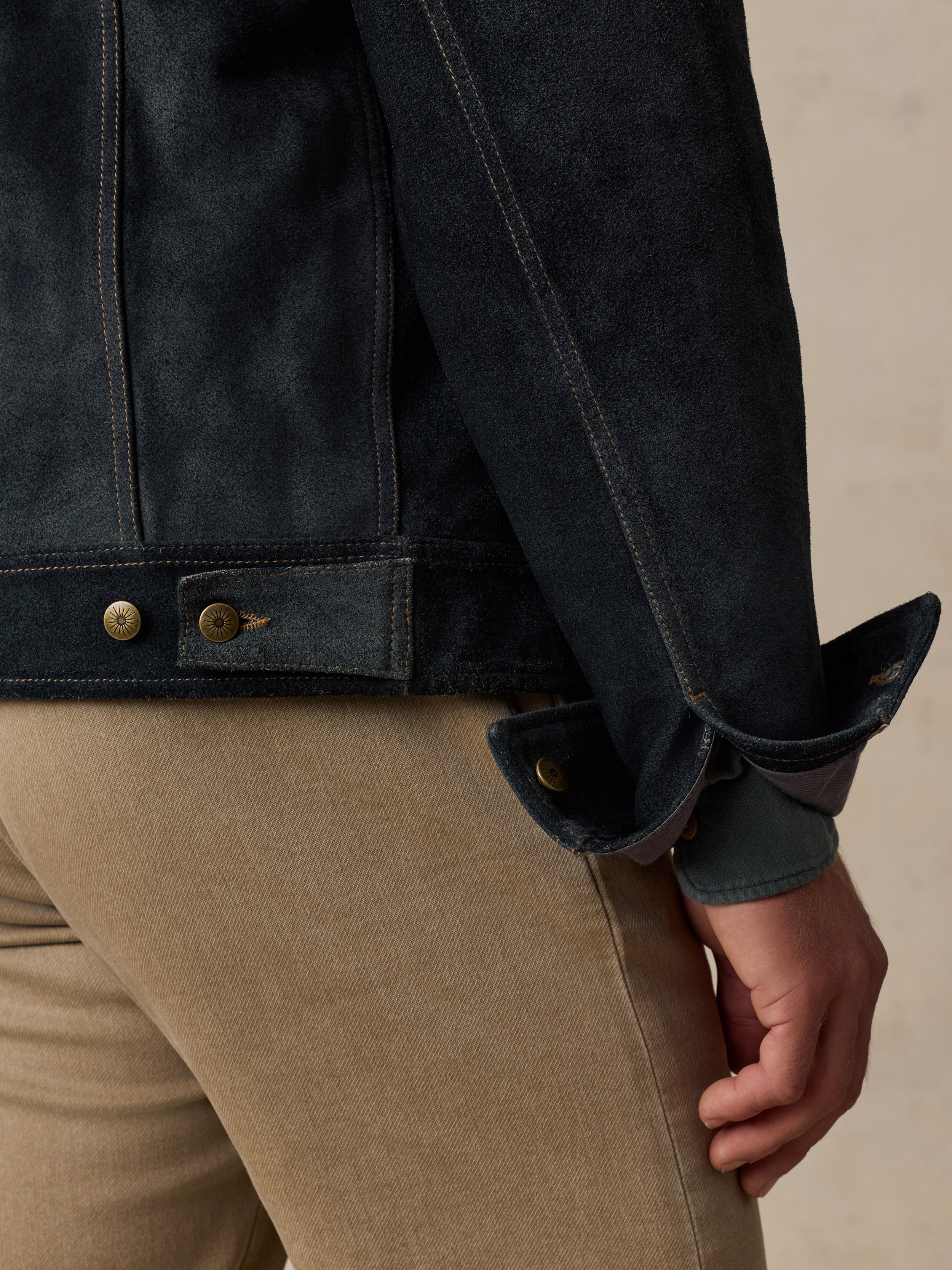 Close-up of a man wearing a dark indigo denim jacket with rolled cuffs and tan pants, showing the sleeve, button details, and hand against a neutral beige background.