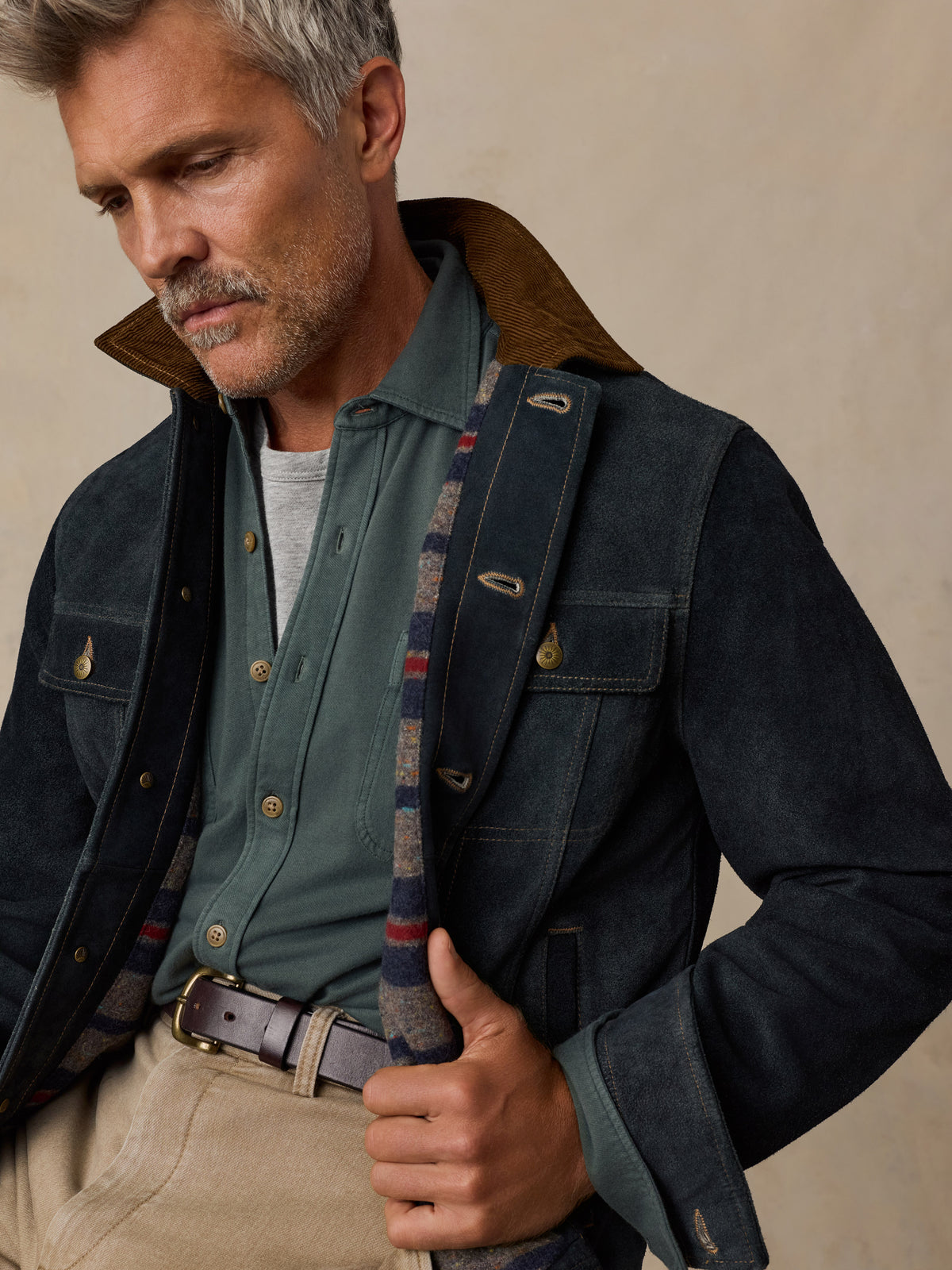 A close-up of a man wearing a dark navy waxed cotton jacket with a brown corduroy collar, layered over a sage green button-down shirt and beige trousers.
