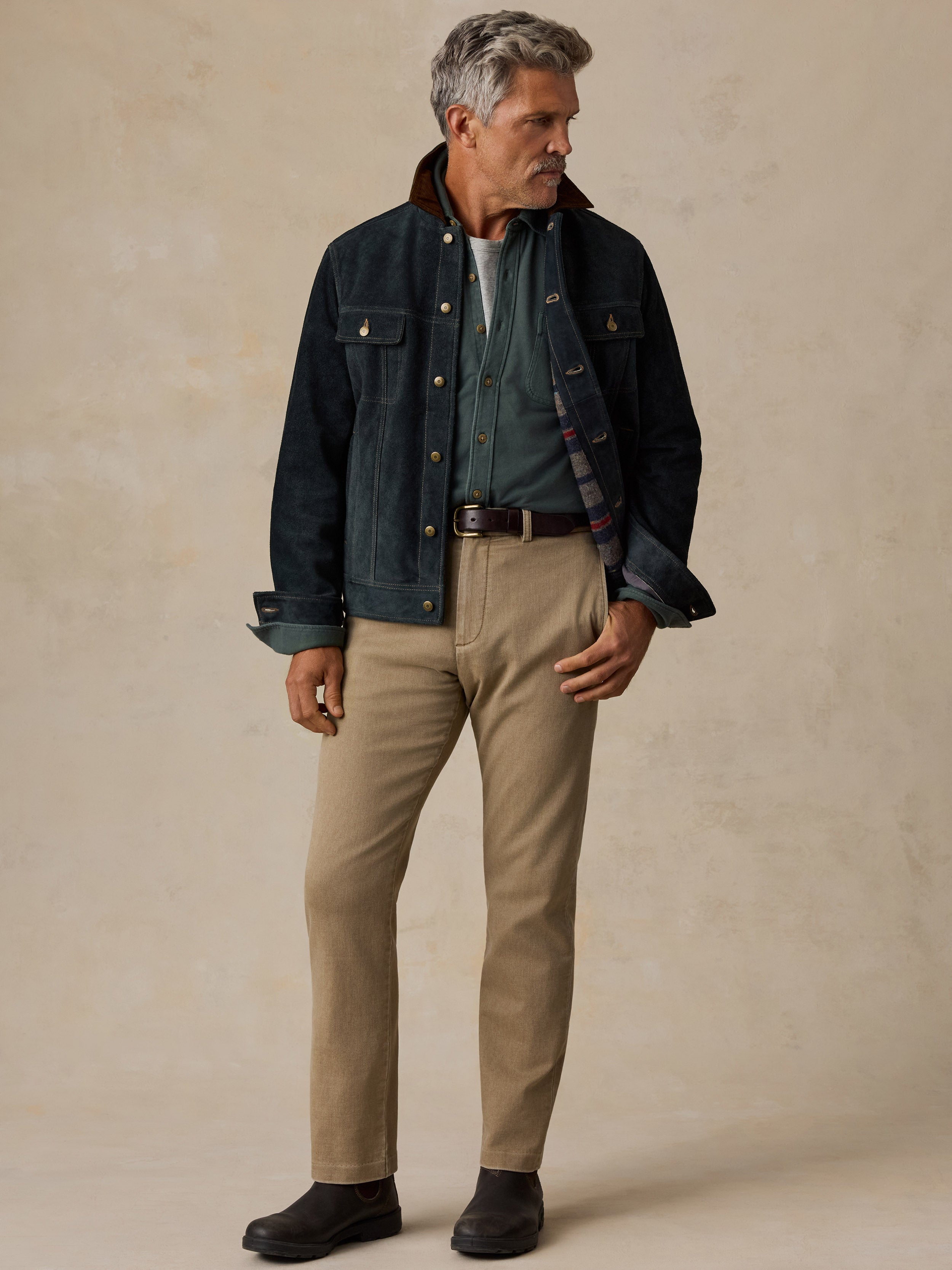 A man stands against a textured beige studio backdrop wearing a dark indigo denim jacket with a brown corduroy collar, a green shirt, khaki pants, and dark brown boots.