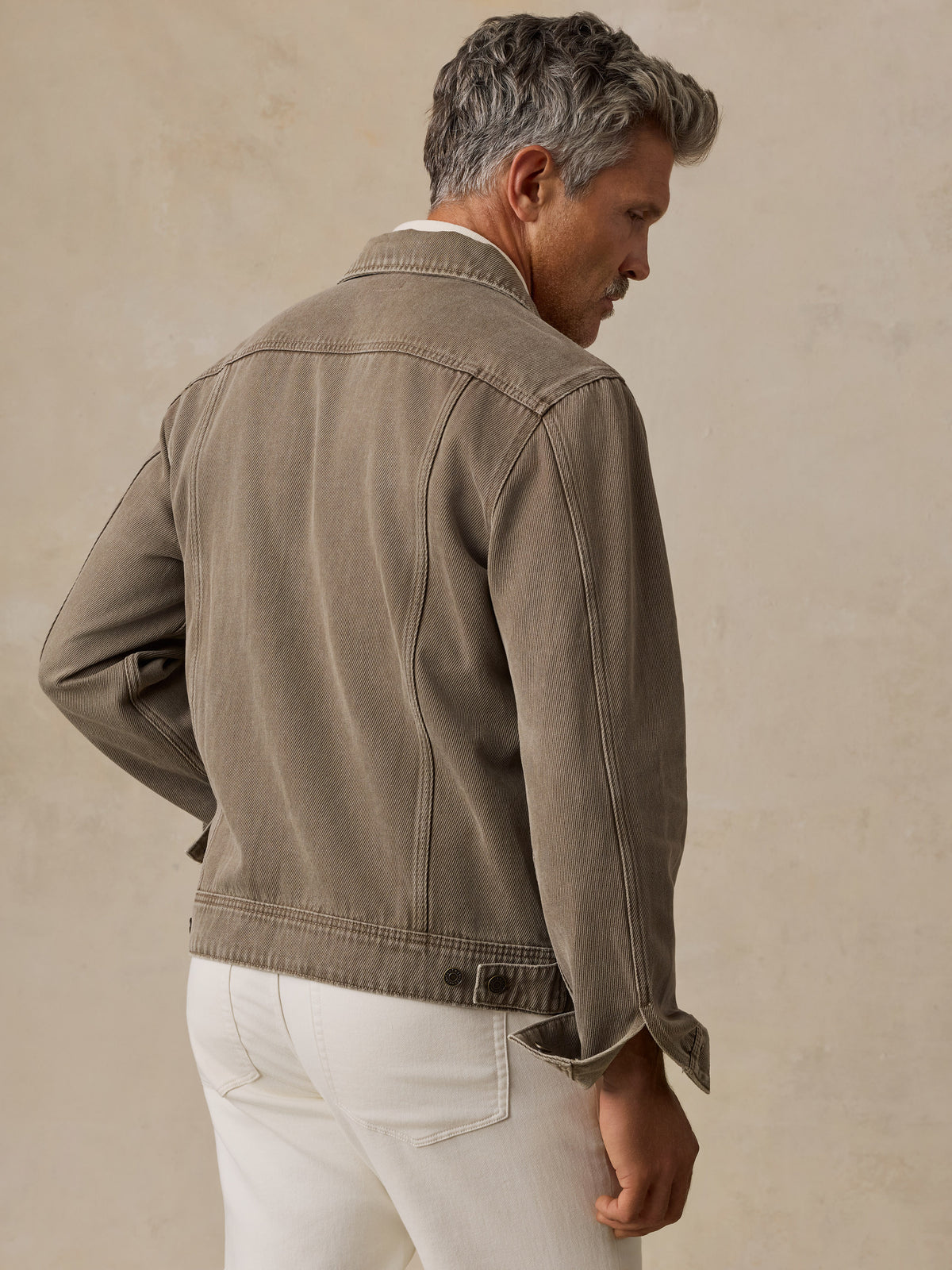 A man stands against a textured beige studio backdrop wearing a taupe jacket over a cream shirt and white trousers