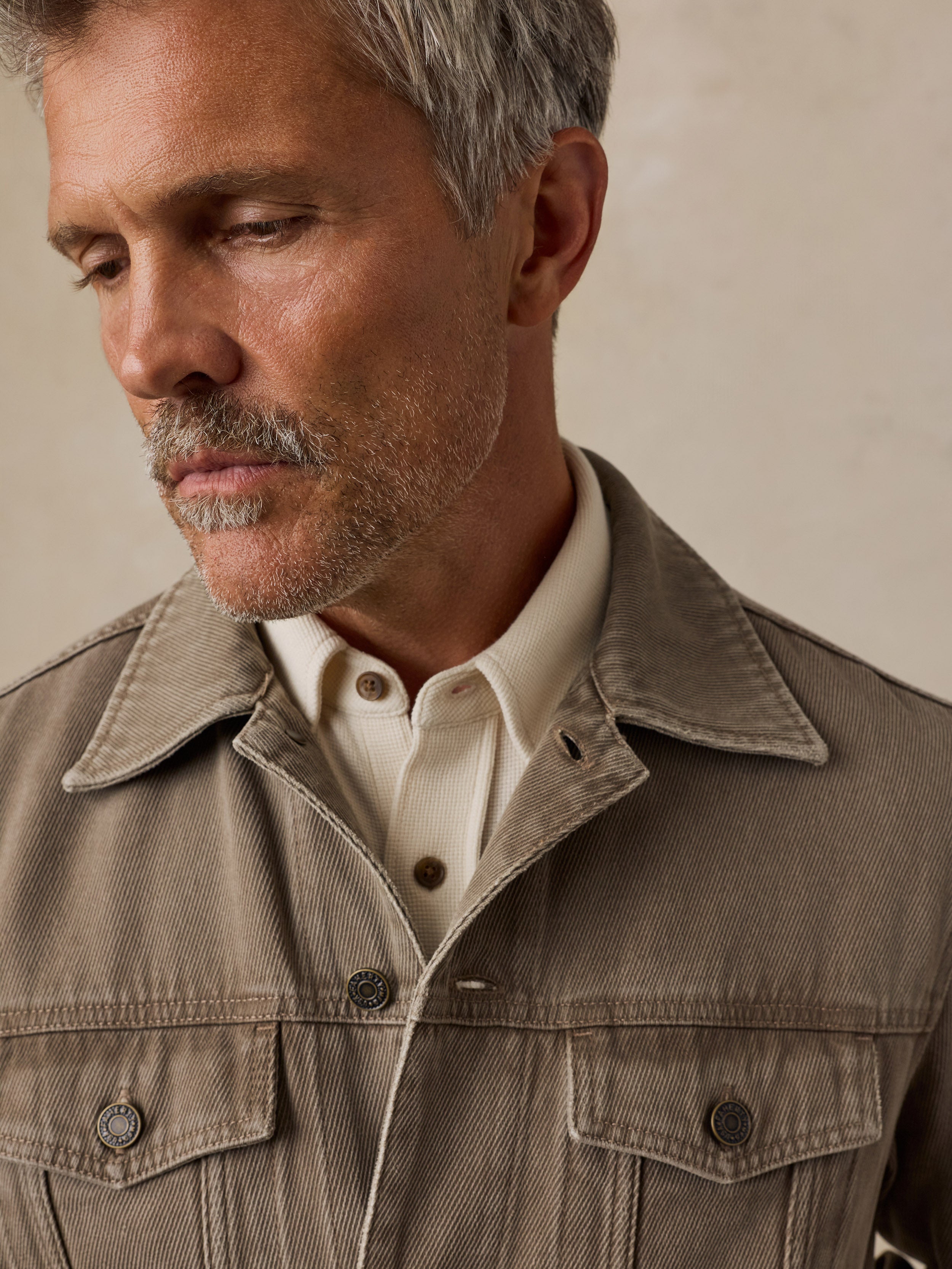 A man stands against a textured beige studio backdrop wearing a taupe jacket over a cream shirt
