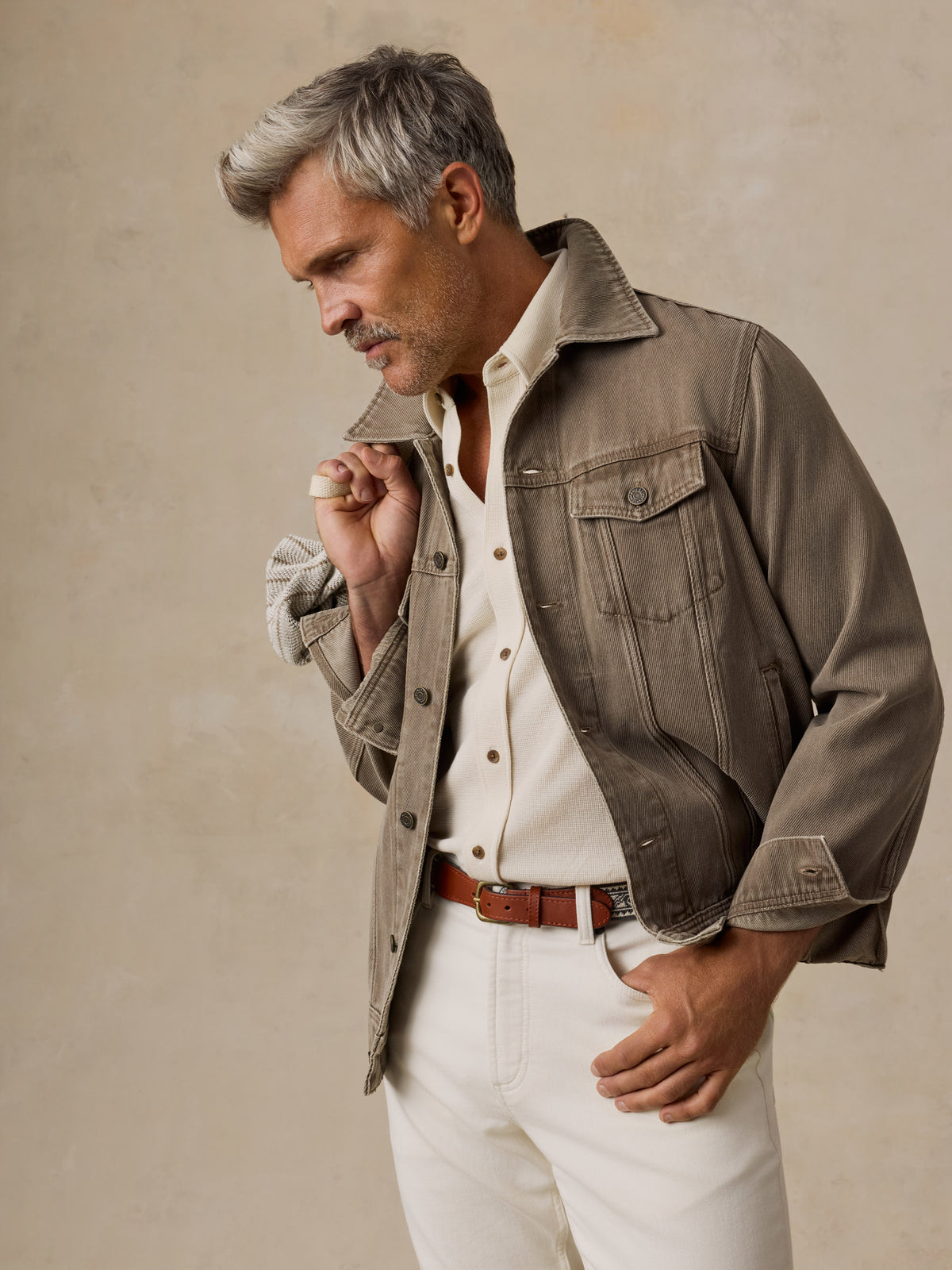 A man stands against a textured beige studio backdrop wearing a taupe jacket over a cream shirt and white trousers