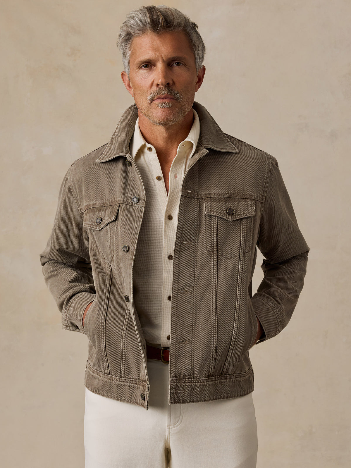 A man stands against a textured beige studio backdrop wearing a taupe jacket over a cream shirt and white trousers