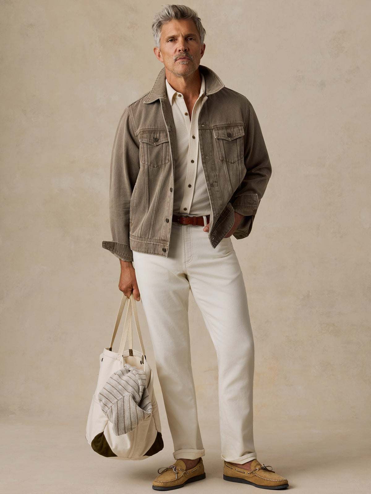 A man stands against a textured beige studio backdrop wearing a taupe jacket over a cream shirt, white trousers, tan loafers, and holding a cream tote bag.