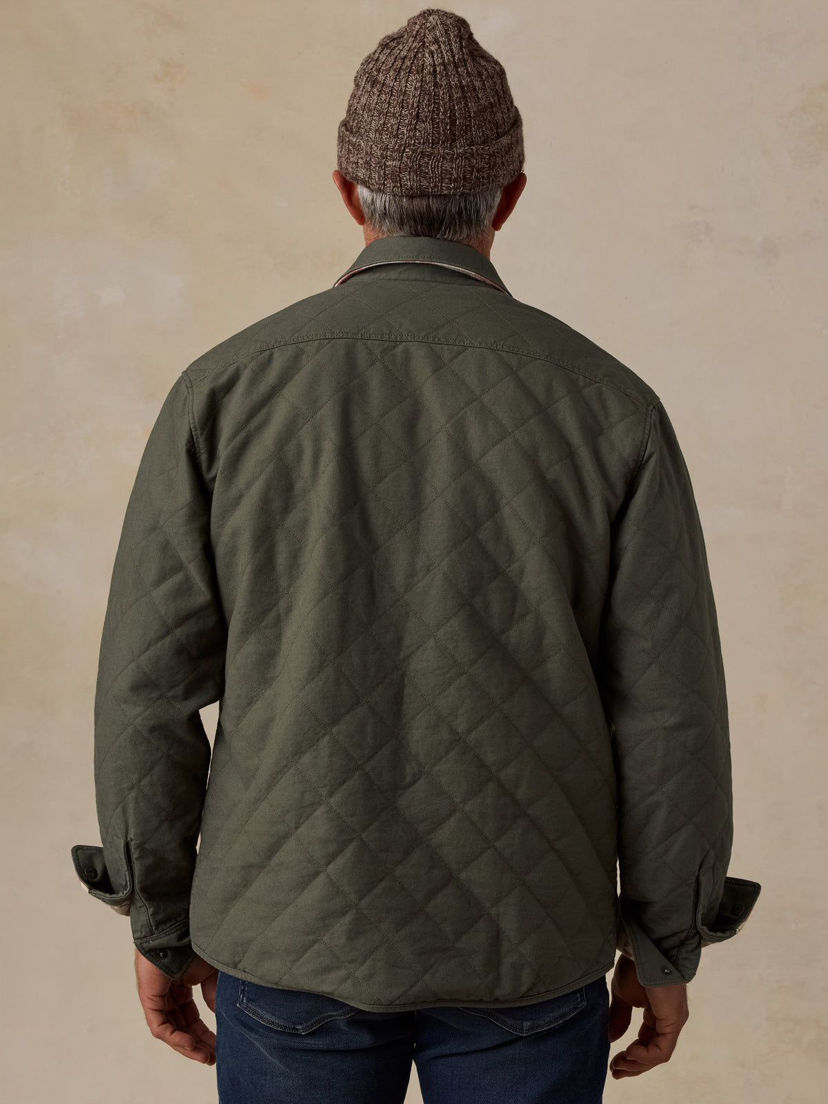 A man viewed from behind wearing an olive green diamond-quilted jacket, a brown knit beanie, and blue jeans, standing against a textured beige wall., image 4 of 5