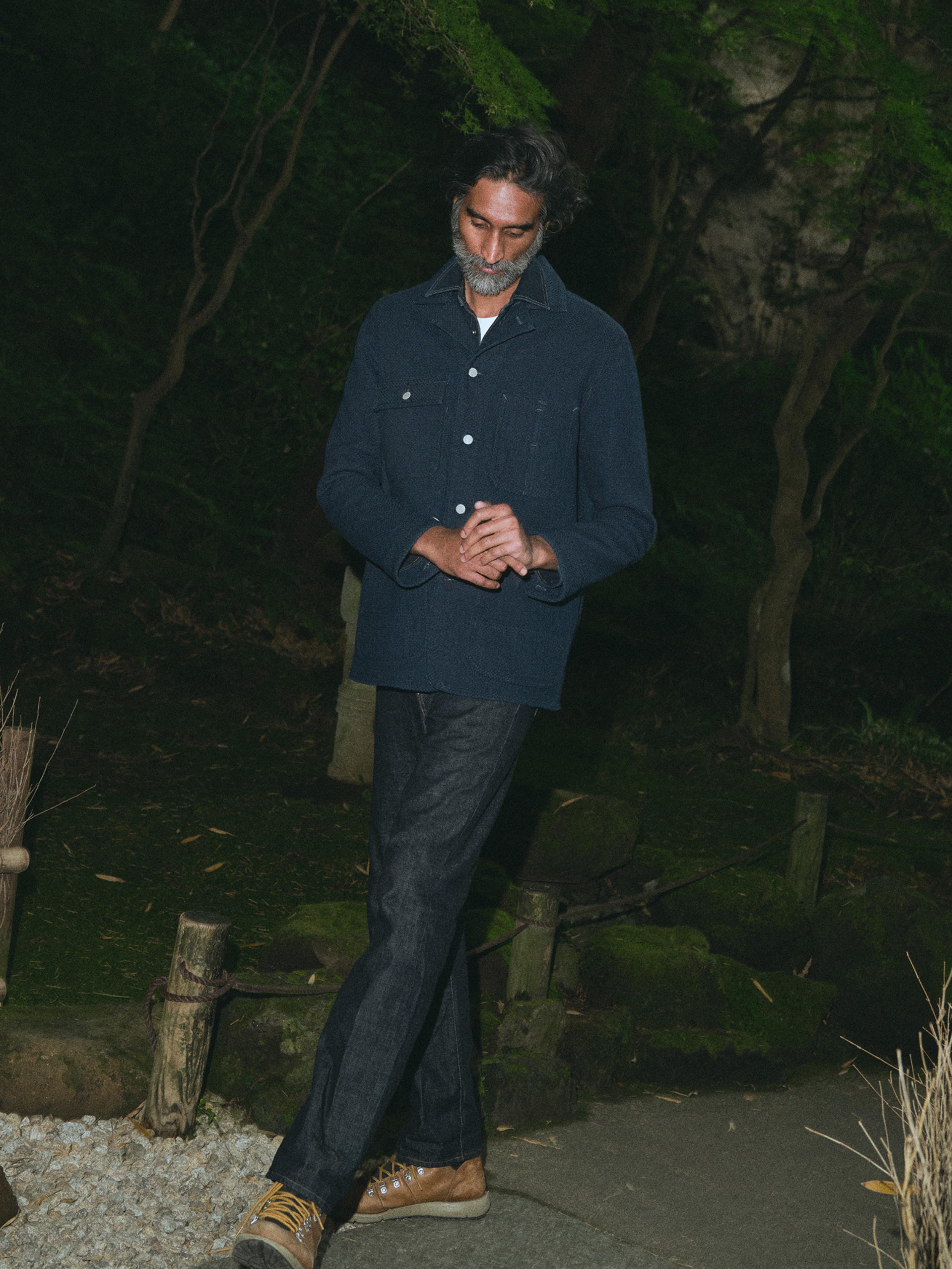 A man with a graying beard wearing a dark indigo denim button-up shirt over a white t-shirt and dark jeans, standing outdoors against a dark, blurry background of foliage.