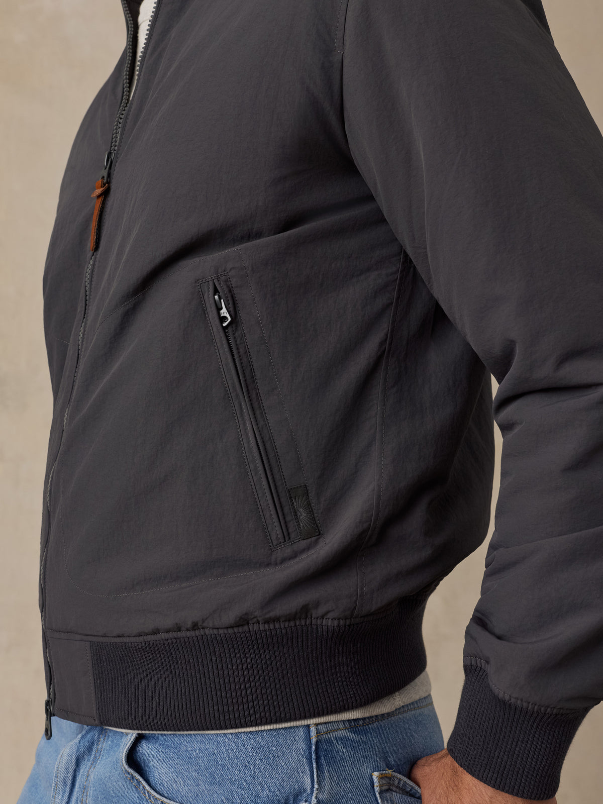 Close-up of a man wearing a dark charcoal zip-front jacket with a zippered chest pocket and ribbed knit collar, cuffs, and hem, paired with blue jeans., image 3 of 6