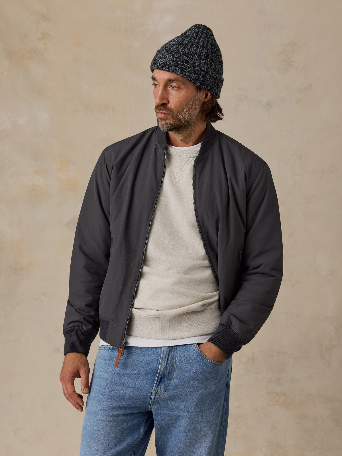 A man wearing a dark charcoal gray bomber jacket, a light gray crewneck sweater, blue jeans, and a dark gray knit beanie, standing against a textured beige studio background., image 6 of 6