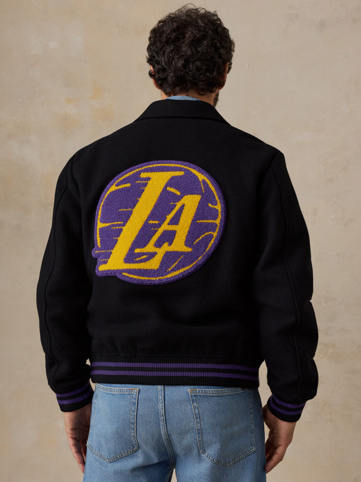 A man viewed from behind wearing a black LA Lakers varsity jacket and blue jeans, image 2 of 8