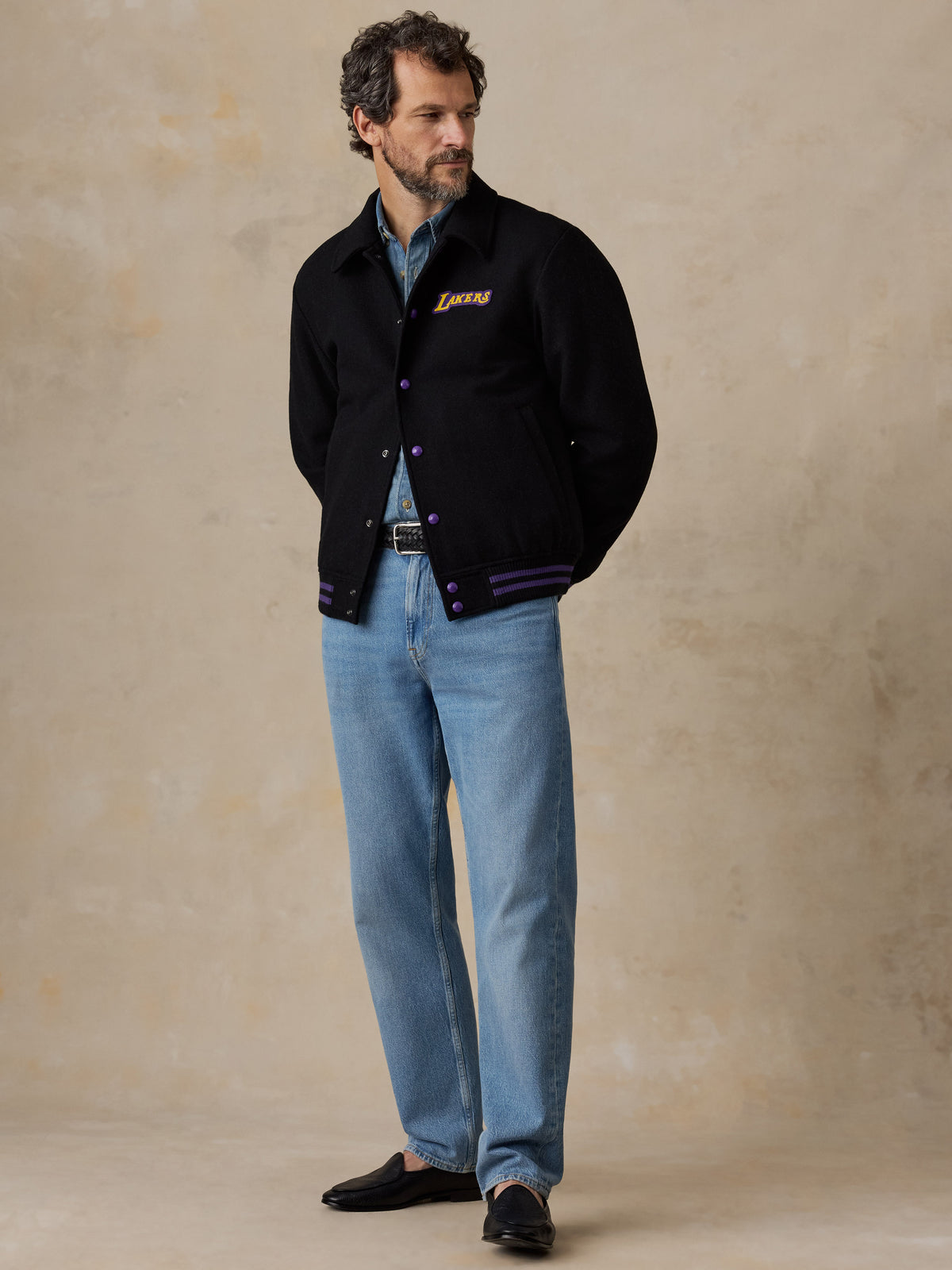A man wearing a black LA Lakers varsity jacket and blue jeans, image 3 of 8
