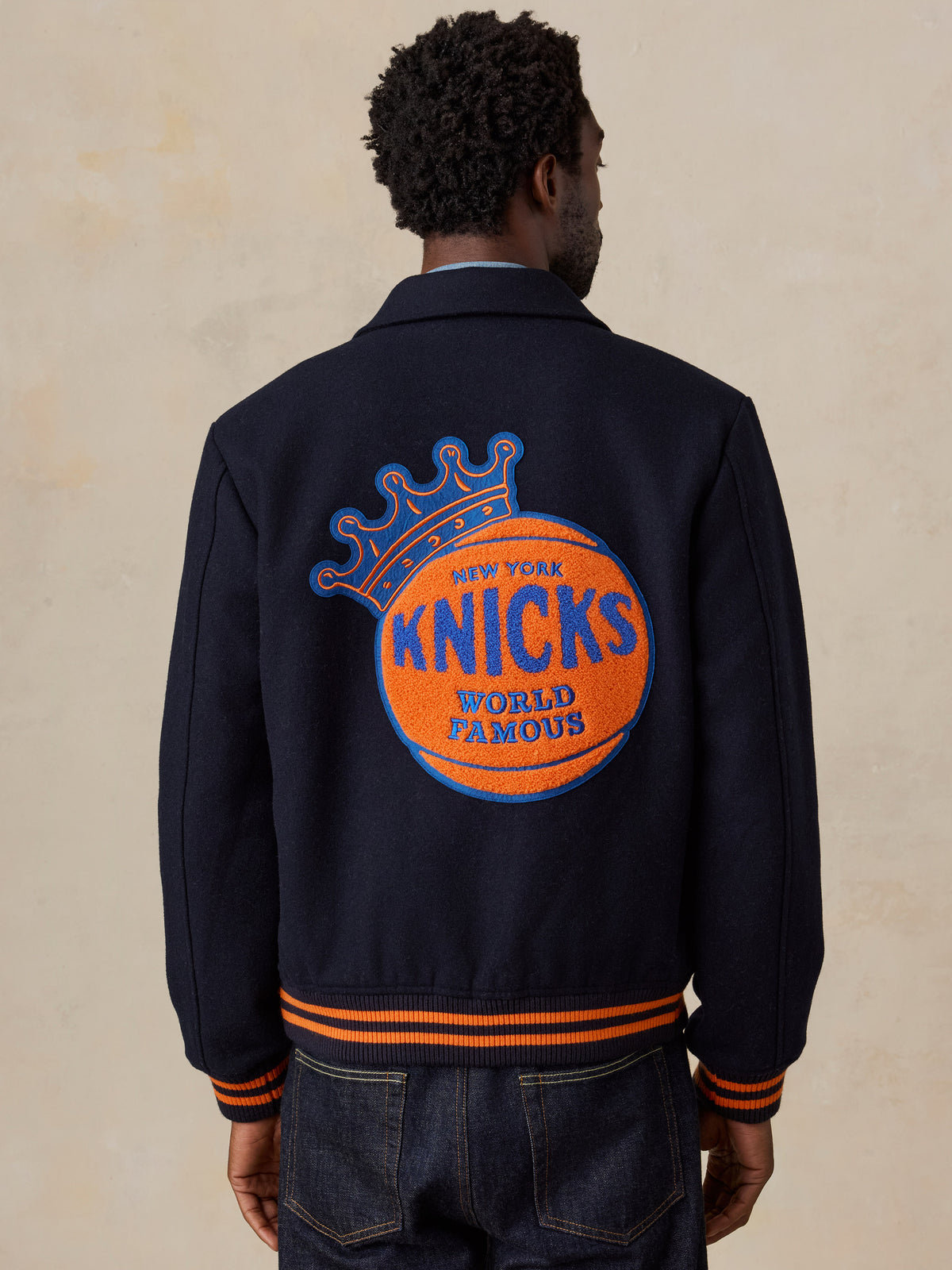 A man viewed from behind wearing a NY Knicks varsity jacket and jeans, image 3 of 8