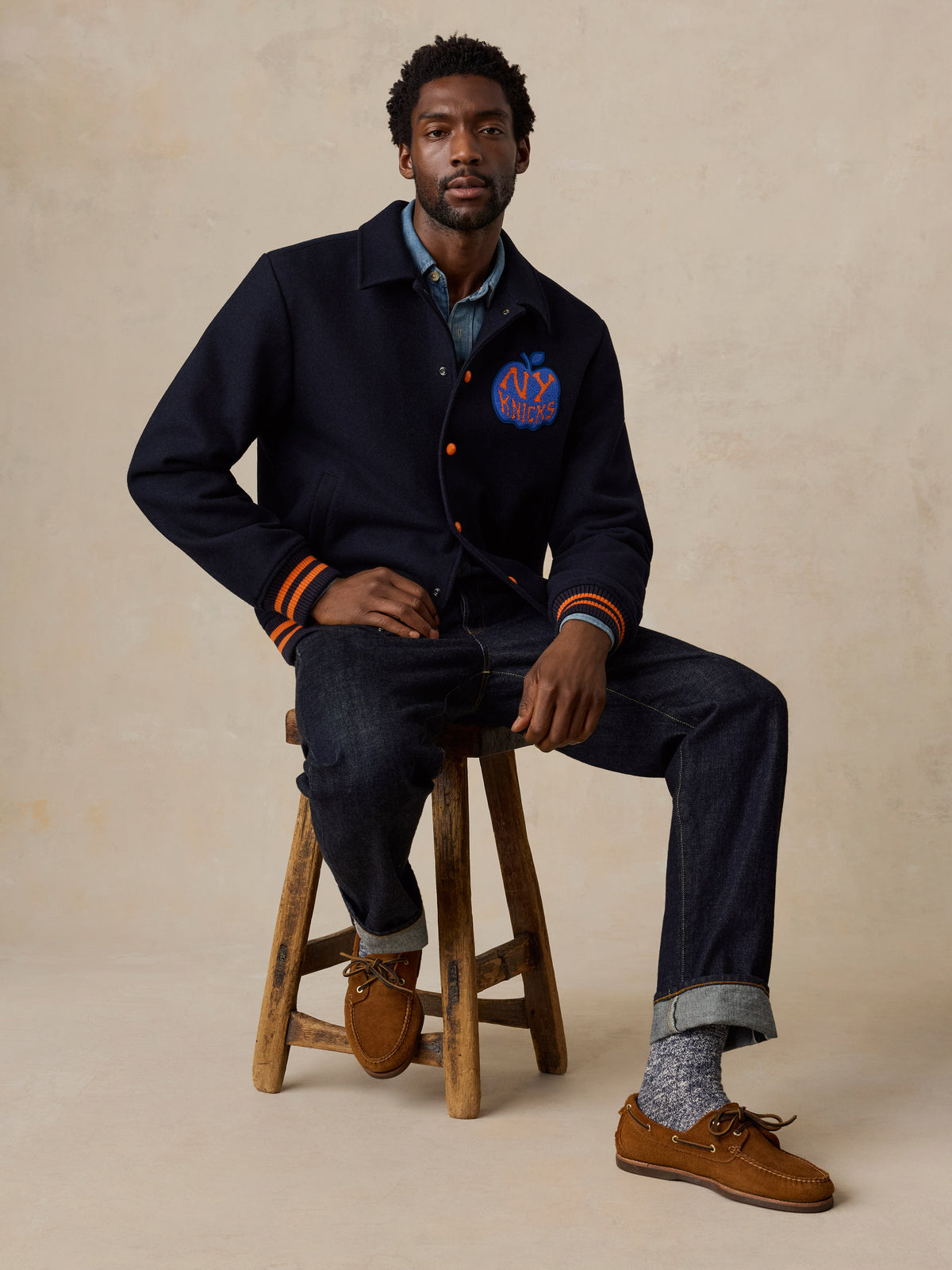 A man wearing a NY Knicks varsity jacket and jeans, image 6 of 8