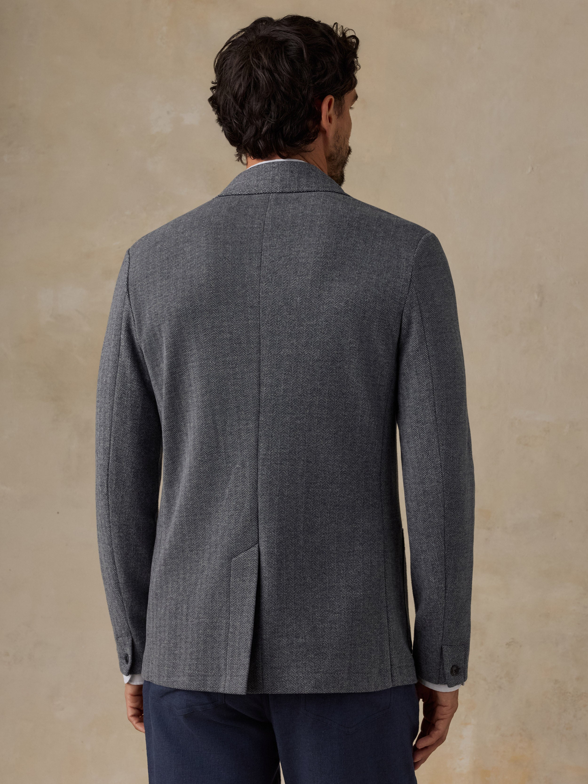 A man viewed from the back wearing a dark gray textured blazer and navy trousers, standing against a warm beige textured wall.