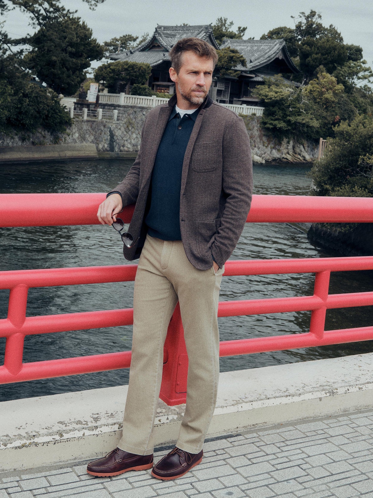 A man stands on a bridge wearing a brown tweed blazer, navy sweater, beige trousers, and brown leather shoes, with a red railing and water in the background., image 2 of 7