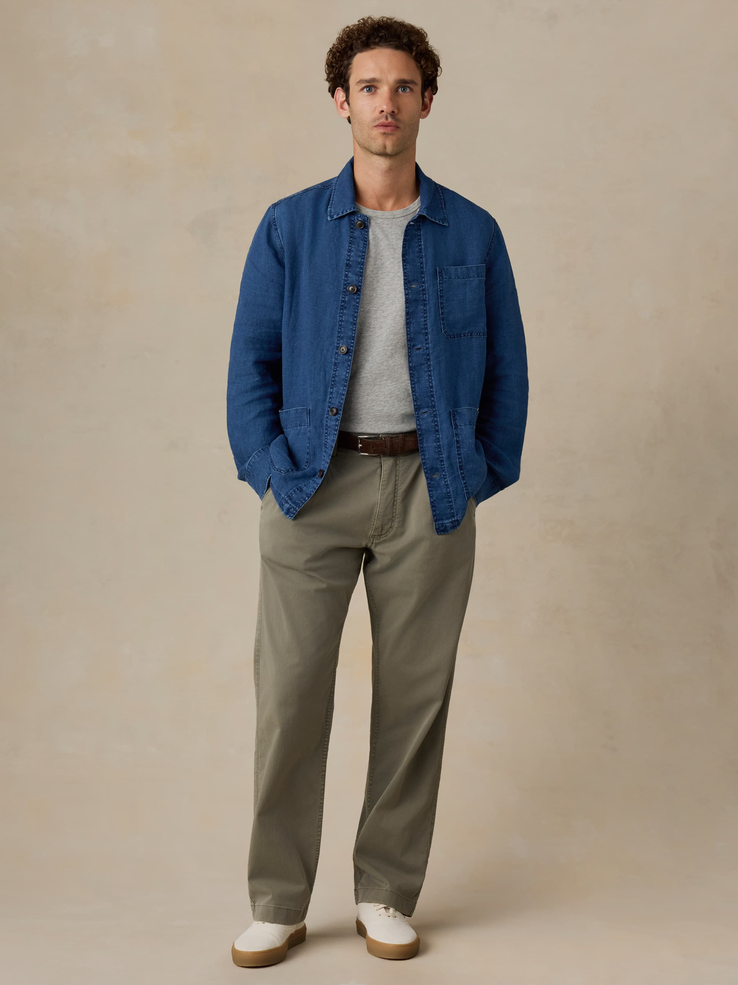 Man wearing a blue denim jacket, gray shirt, and beige pants against a beige background