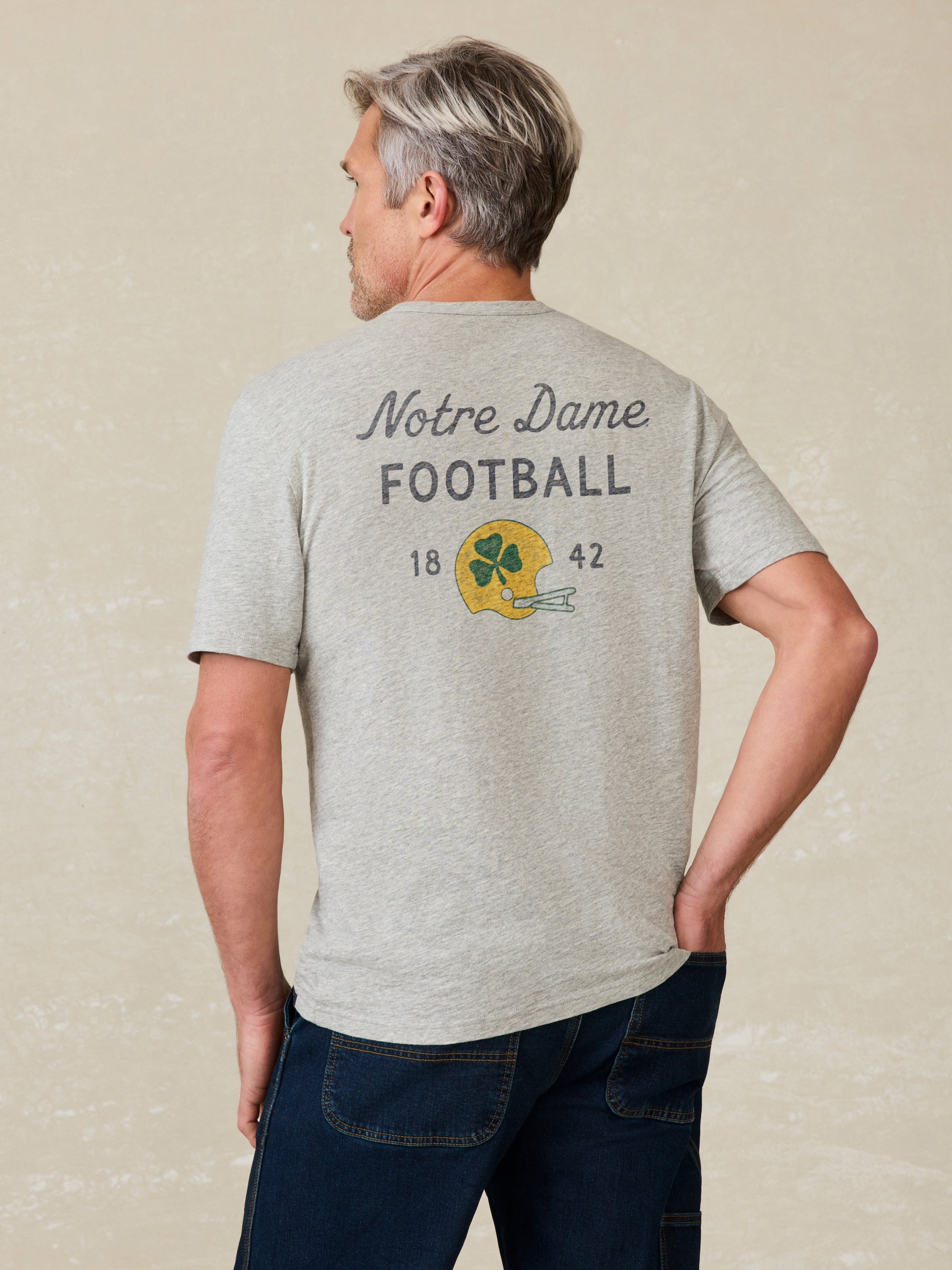 Man wearing a gray t-shirt with 'Notre Dame Football' graphic on the back against a beige background