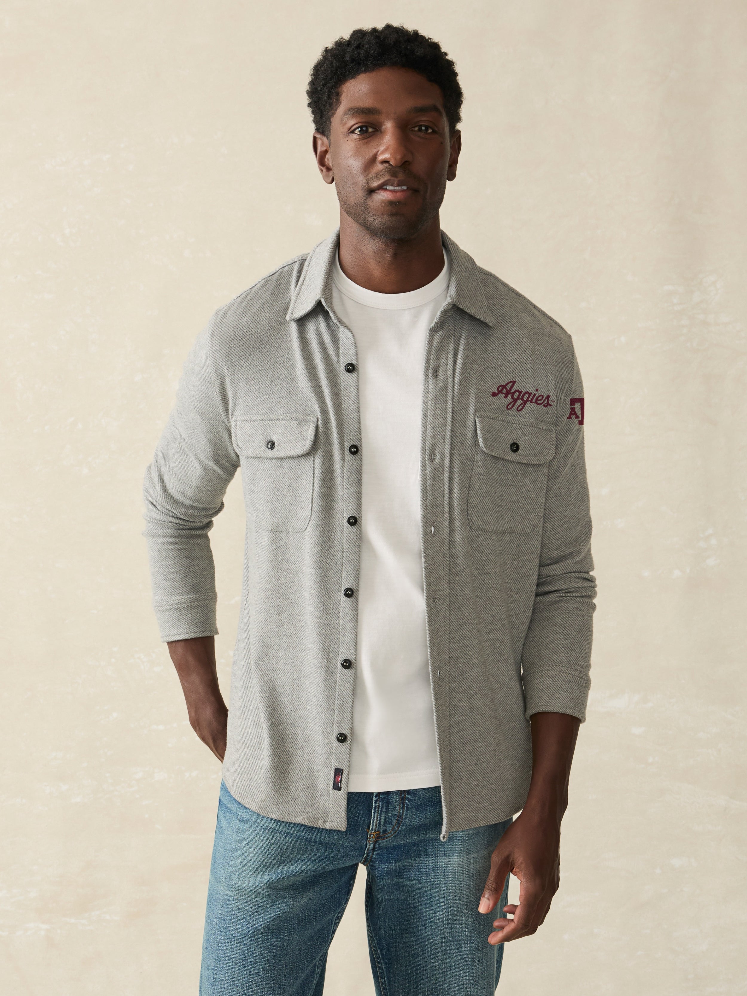 Man is wearing a grey long sleeve shirt with Texas A&M graphic on front left chest and left arm against a beige background
