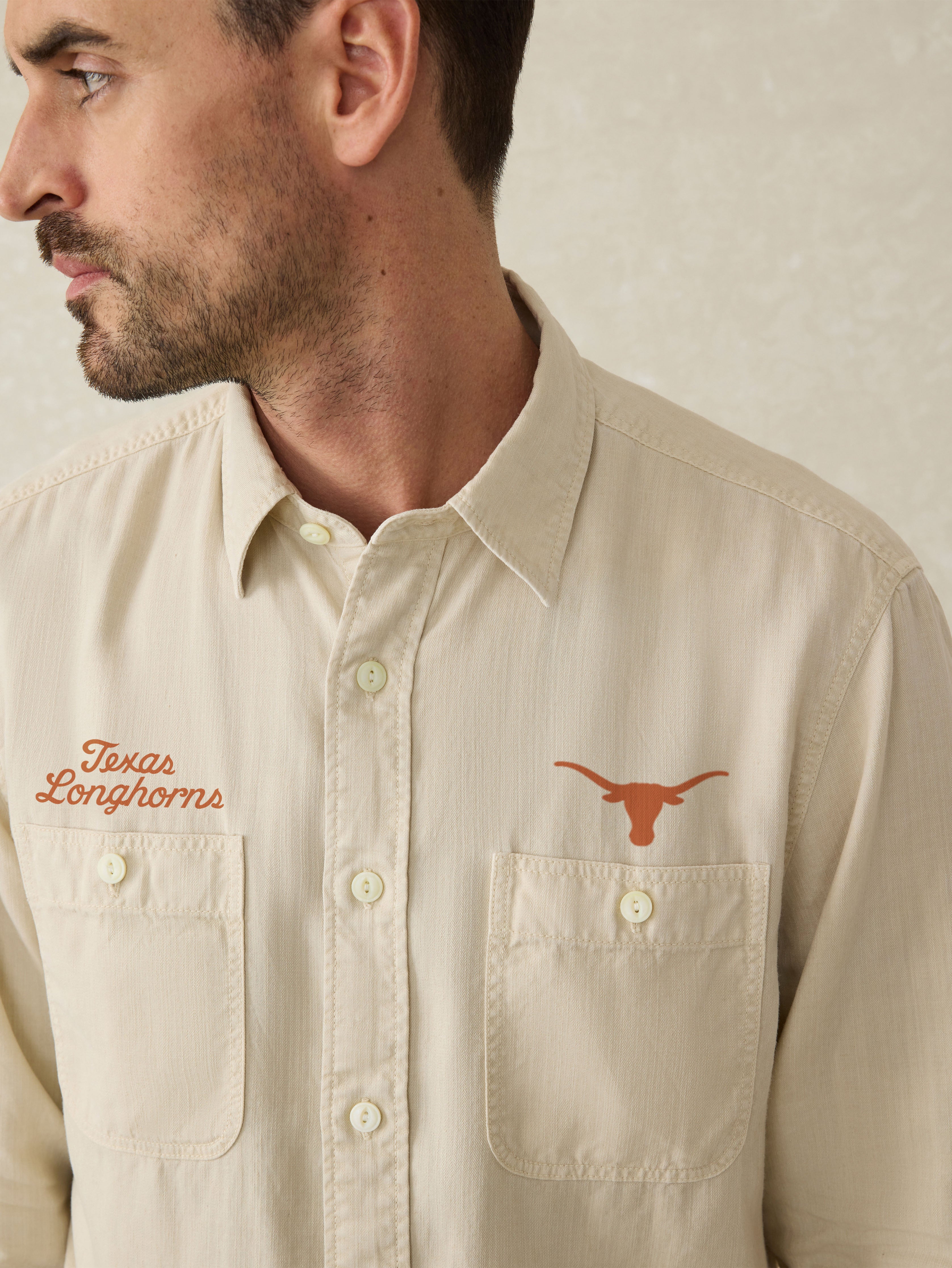 Man wearing an ivory long sleeve button down shirt with an University of Texas logo above the left and right chest pocket against a beige background
