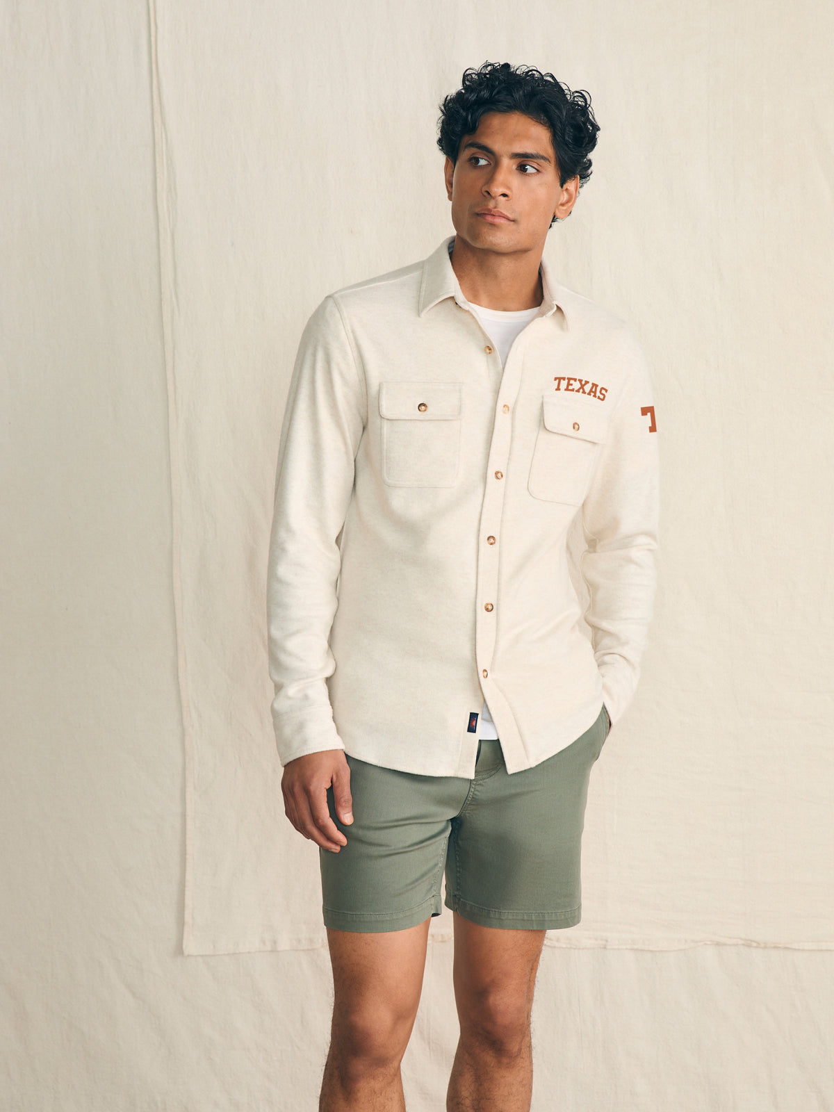 Man wearing a white shirt with UT Austin graphic on the left chest and shoulder against a beige background, image 1 of 3