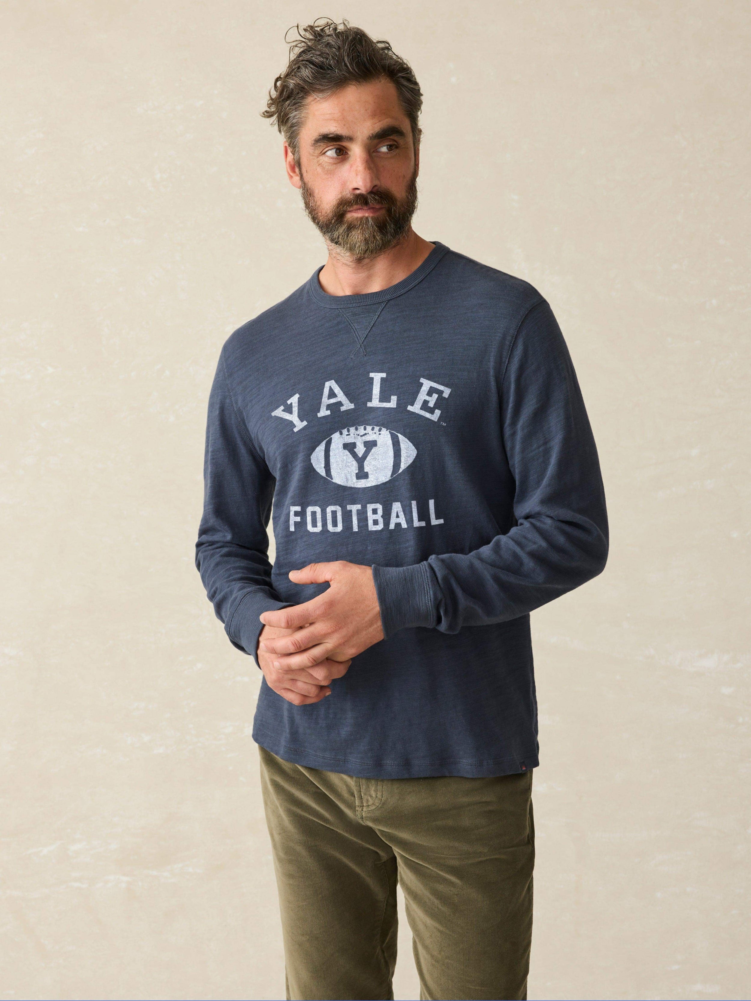 Man is wearing a navy long sleeve shirt with Yale graphic on chest against a beige background