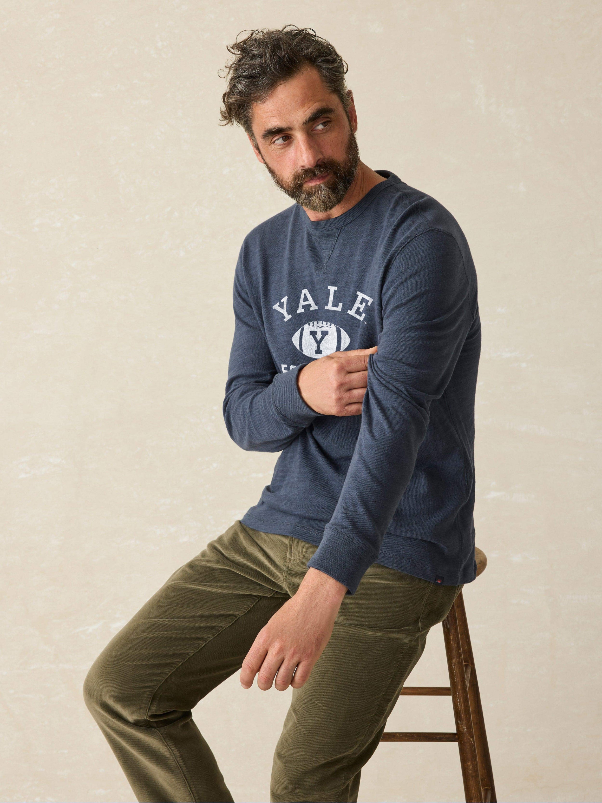 Man is wearing a navy long sleeve shirt with Yale graphic on chest against a beige background
