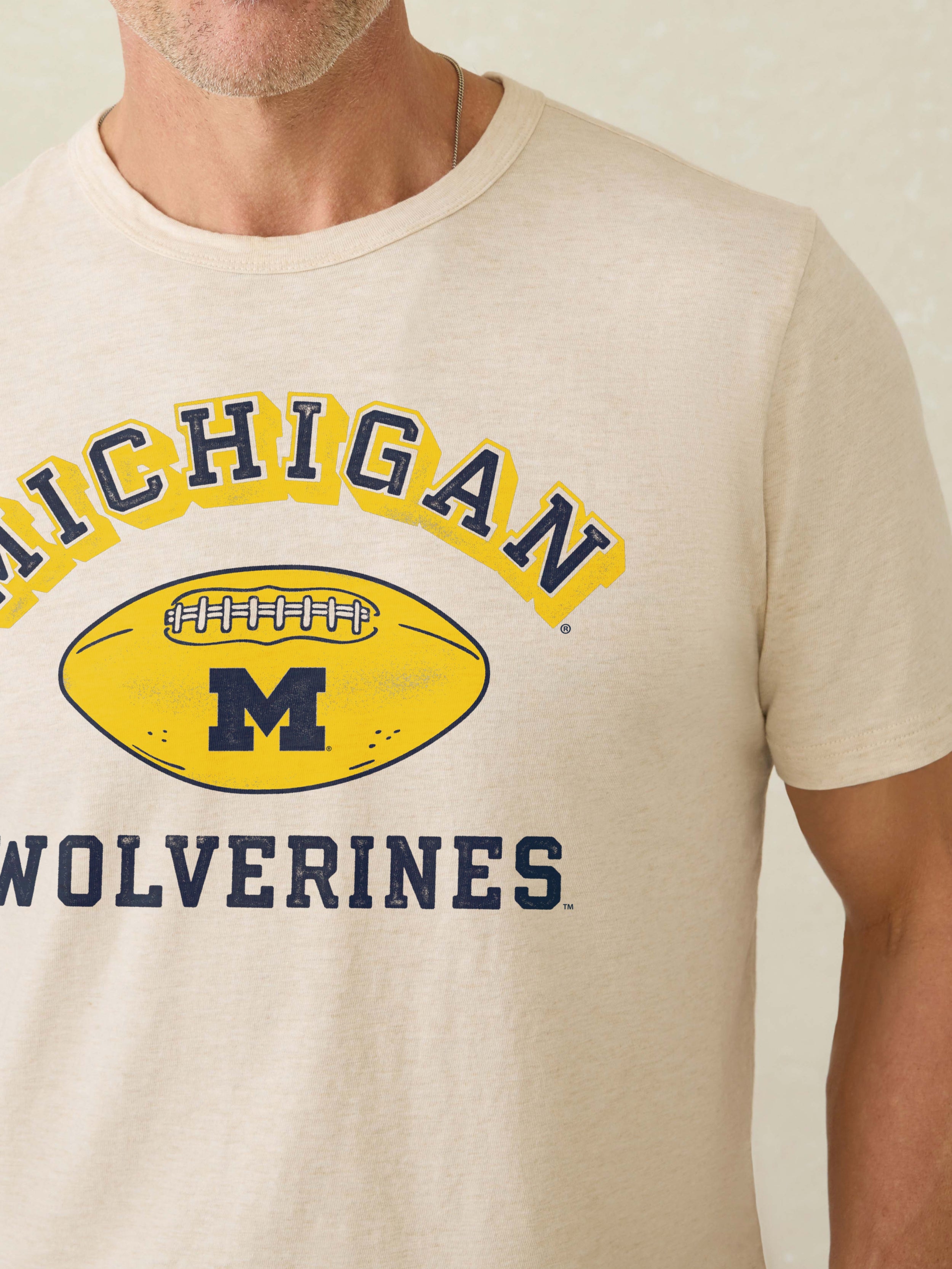 Man wearing a grey short sleeve t-shirt with University of Michigan graphic at the center chest against a beige background