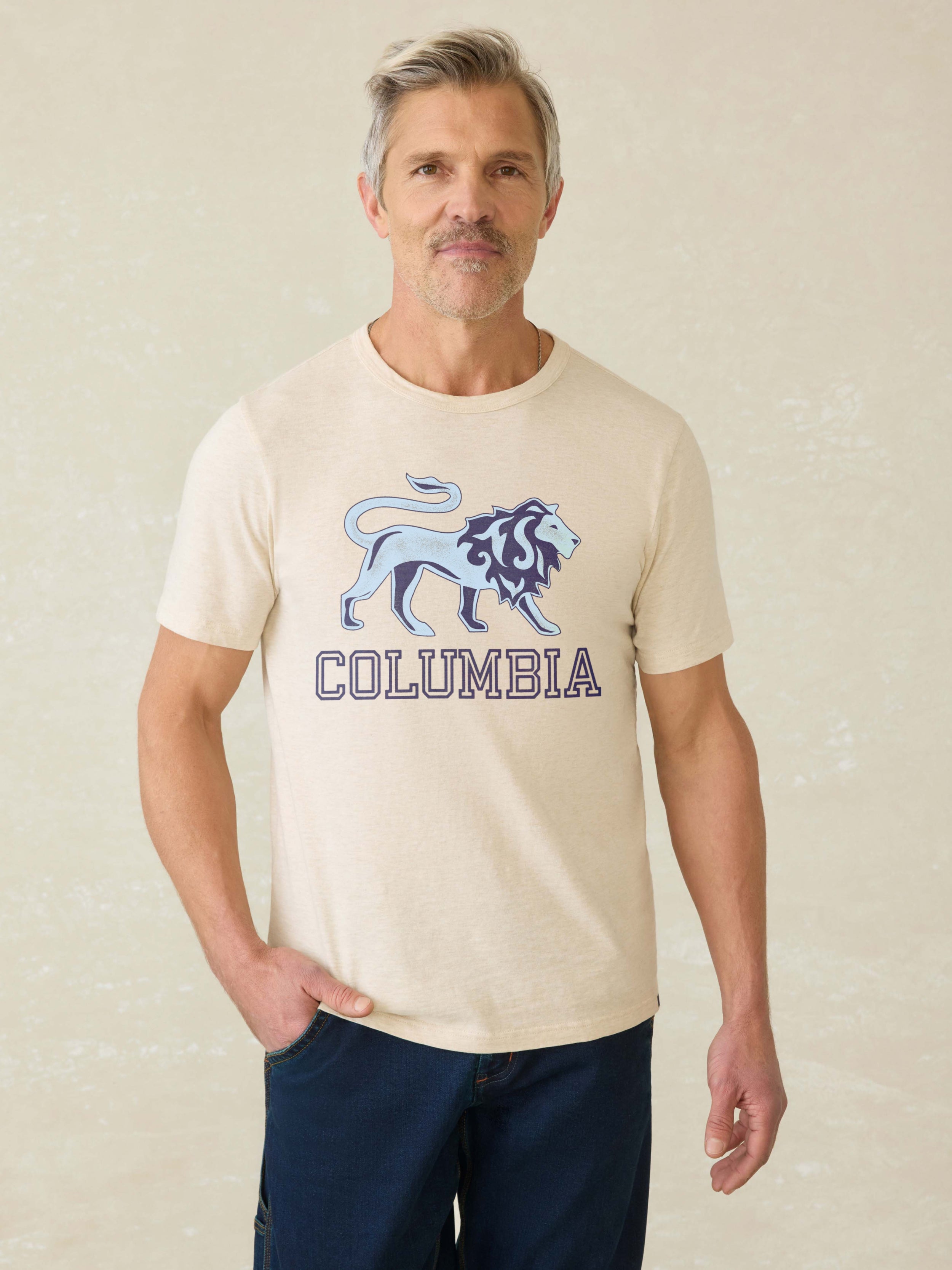 Man wearing a ivory heather t-shirt with Columbia graphic at the front against a beige background