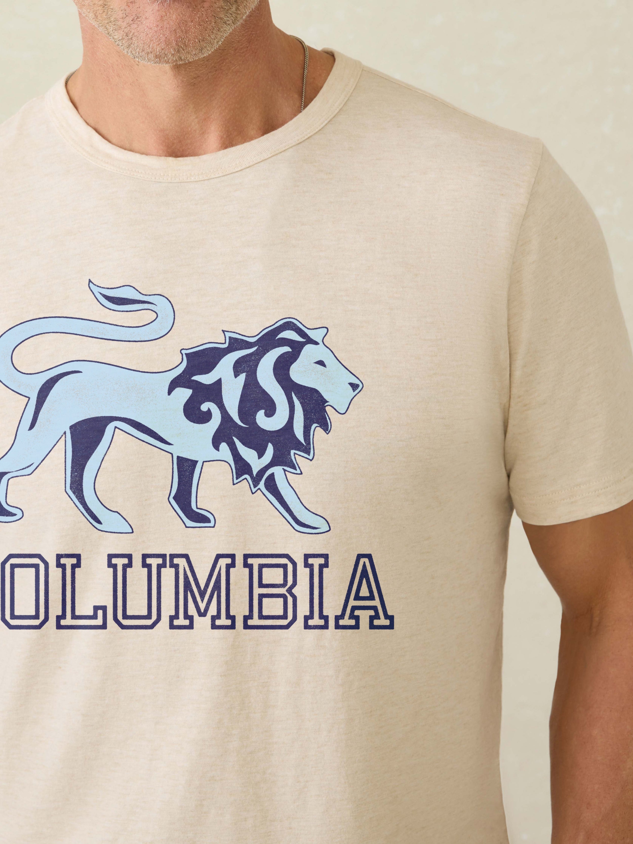 Man wearing a ivory heather t-shirt with Columbia graphic at the front against a beige background