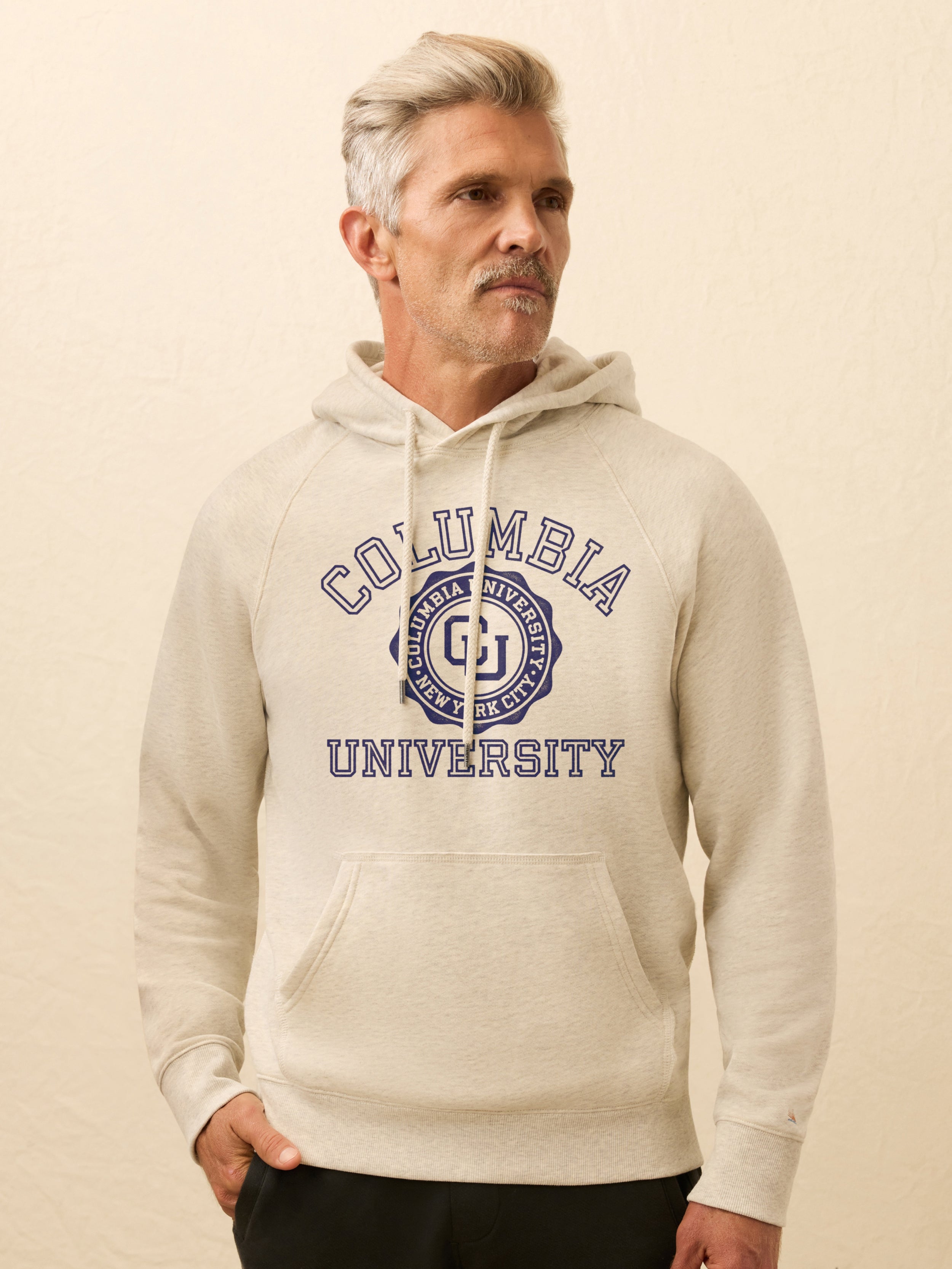 Man is wearing a heather grey hoodie with Columbia graphic on the center front against a beige background