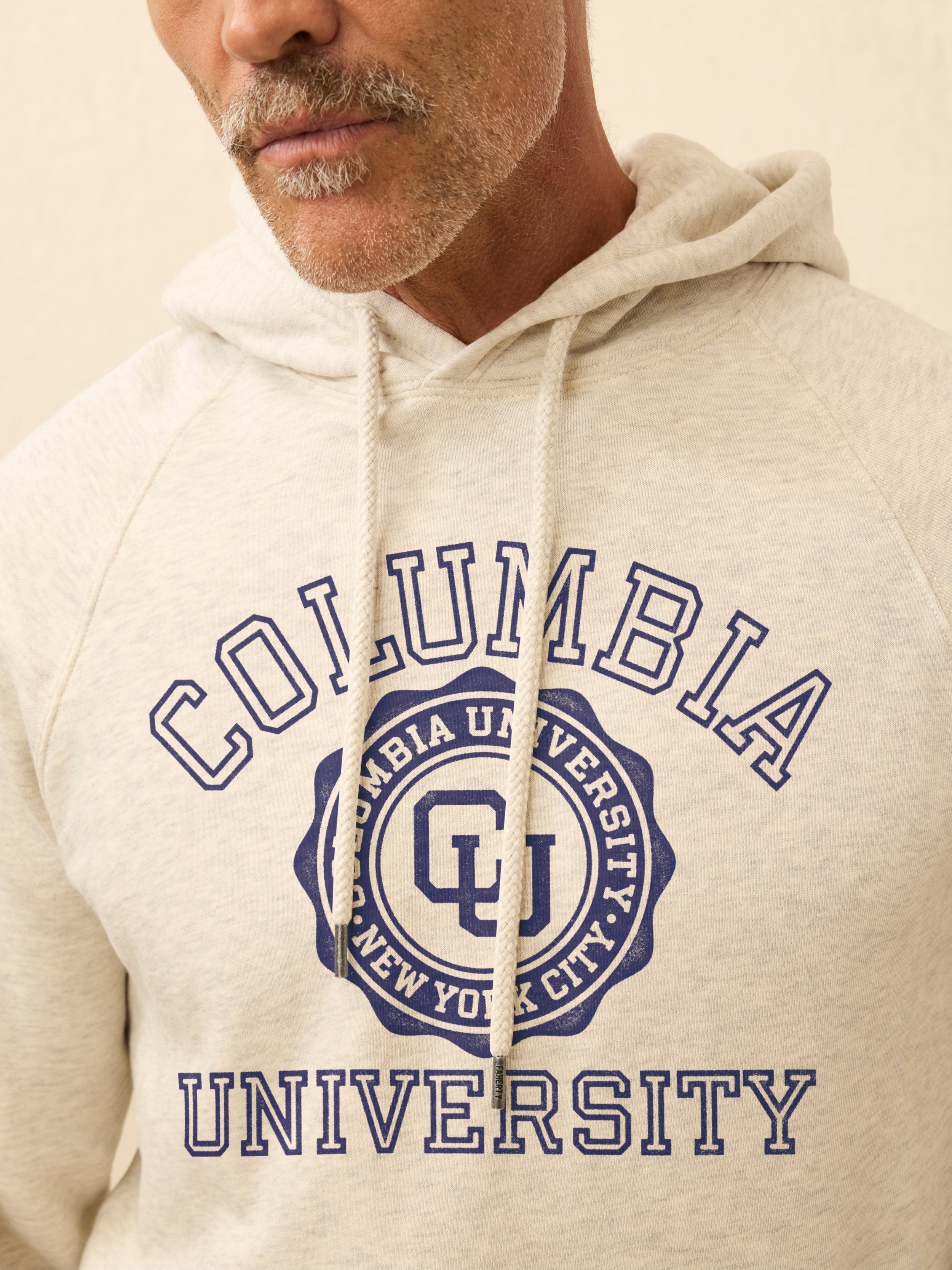 Man is wearing a heather grey hoodie with Columbia graphic on the center front against a beige background