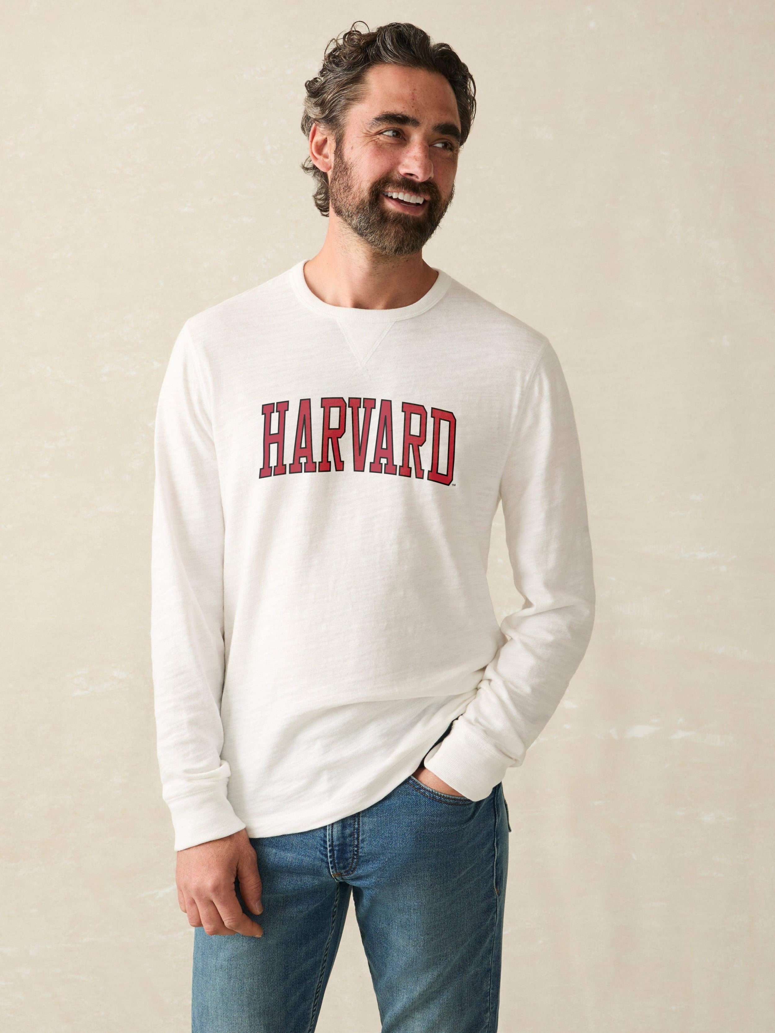 Man wearing a white long sleeve shirt with Harvard graphic at the center chest against a beige background