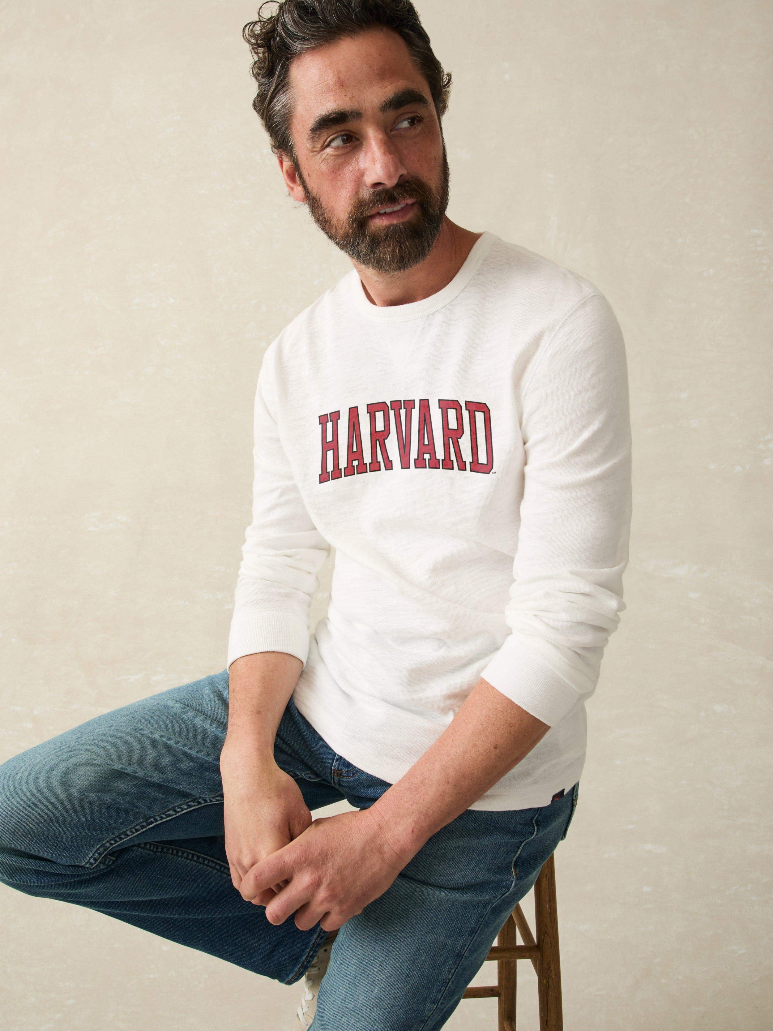Man wearing a white long sleeve shirt with Harvard graphic at the center chest against a beige background