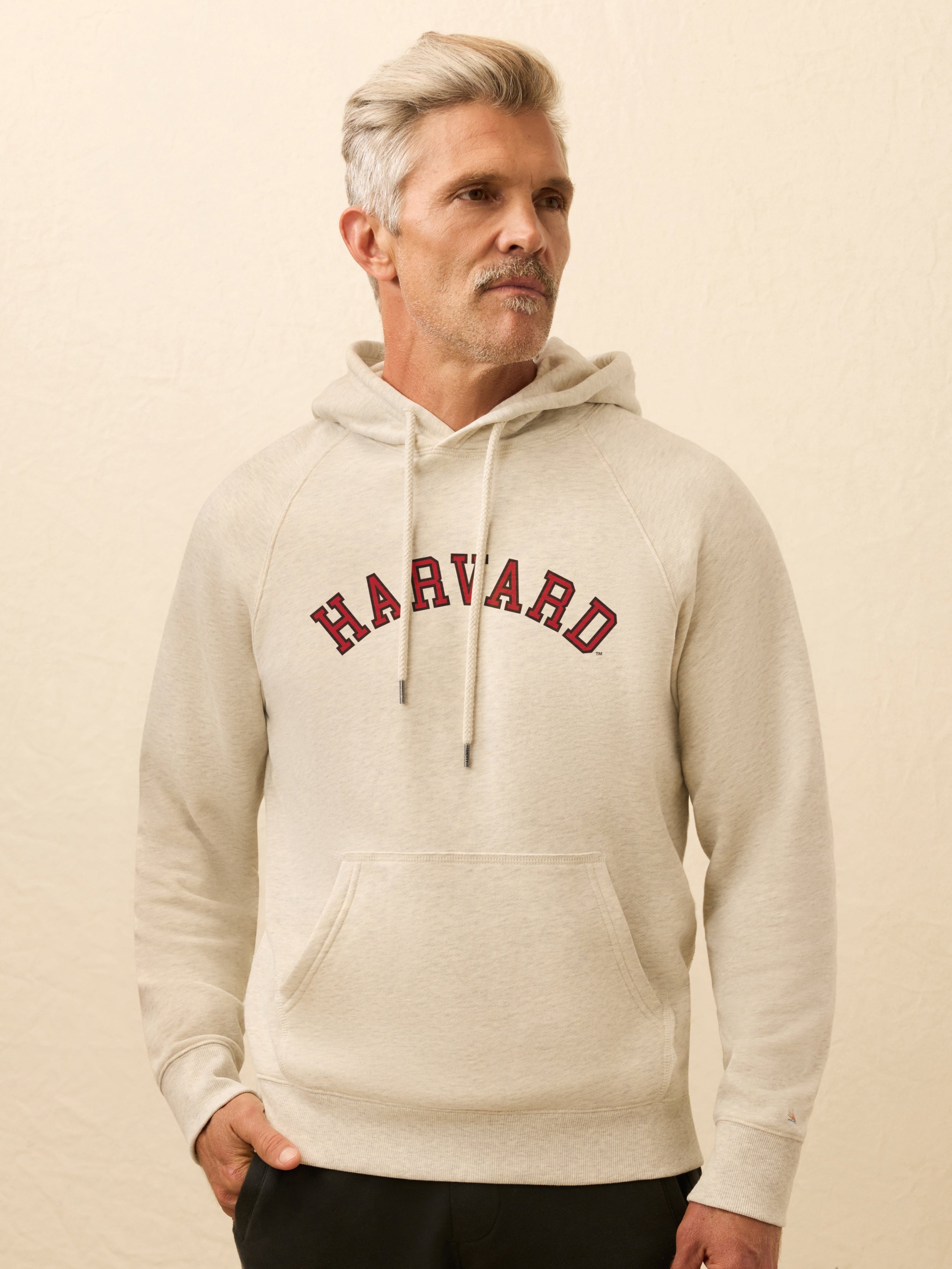 Man wearing a ivory heather hoodie with Harvard graphic on the center front against a beige background