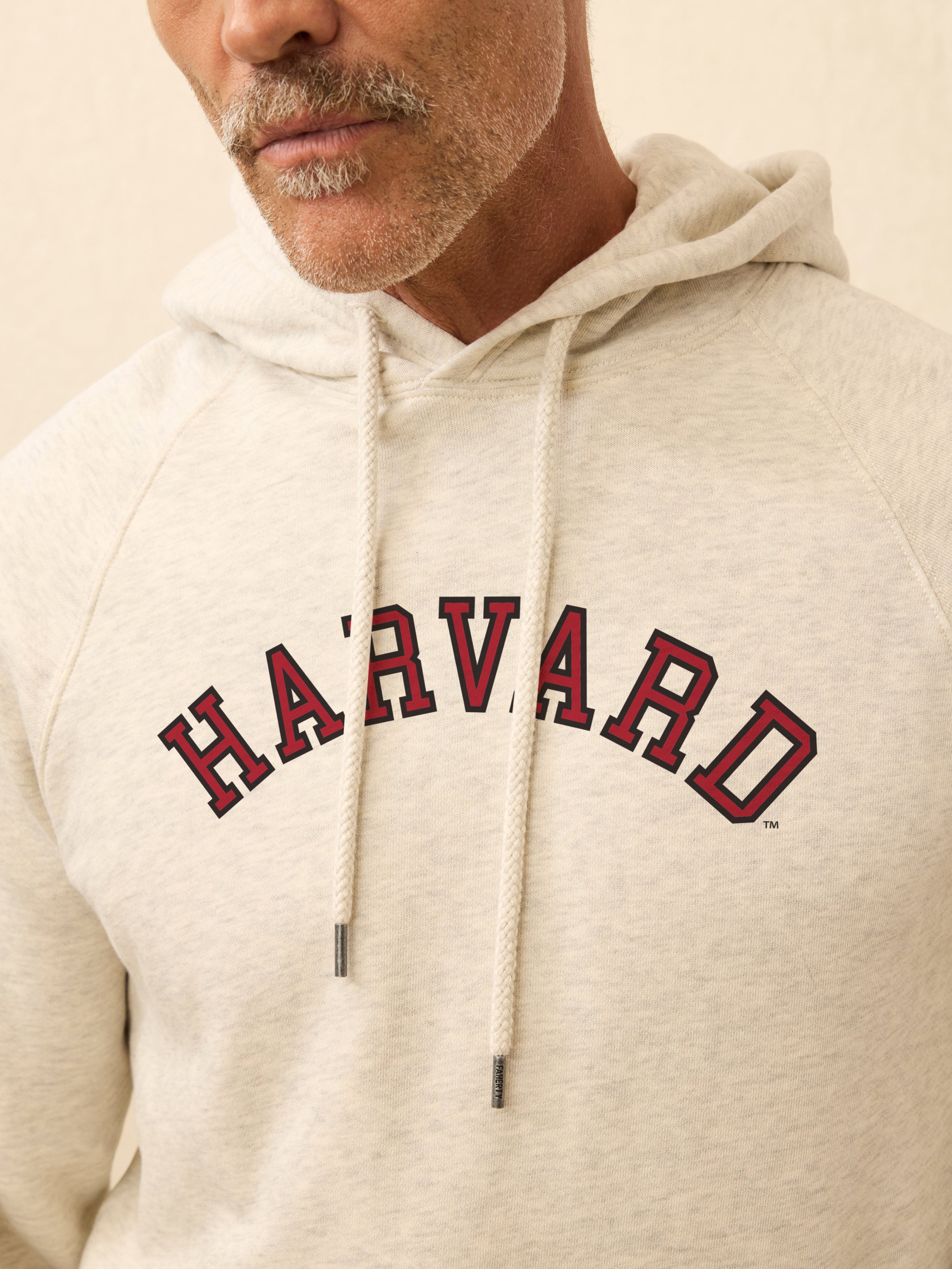 Man wearing a ivory heather hoodie with Harvard graphic on the center front against a beige background