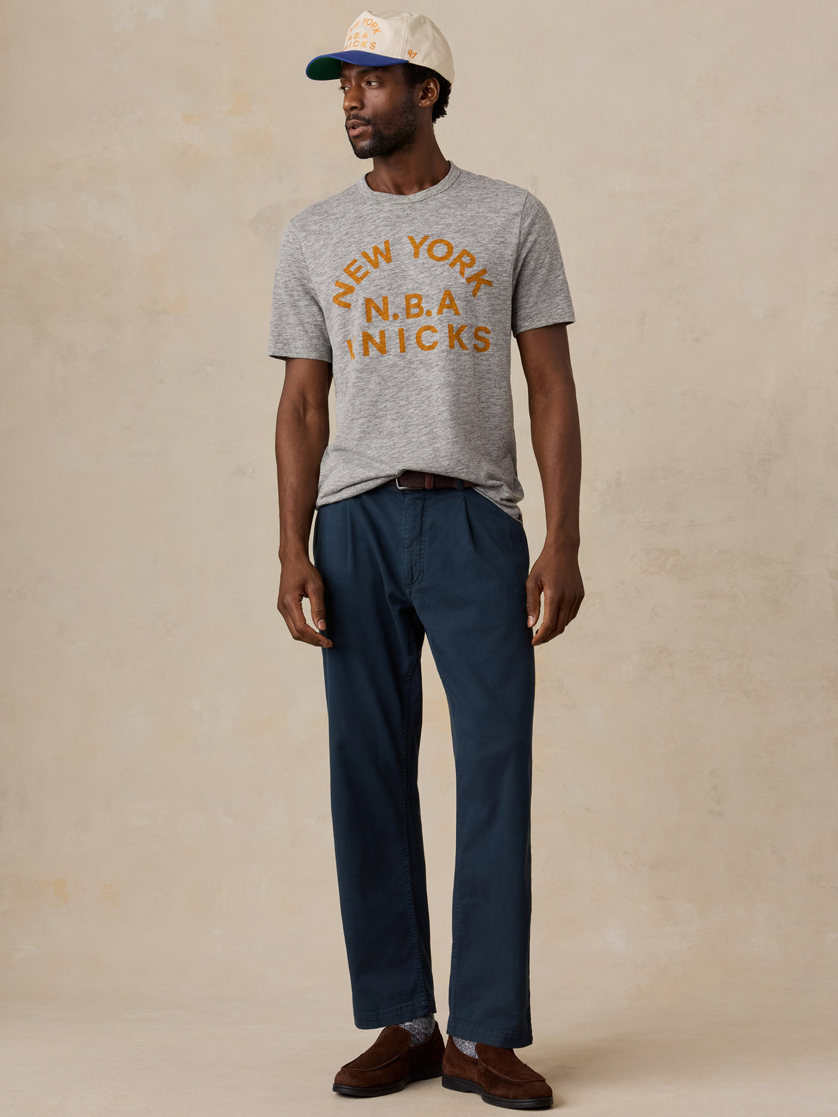 A man wearing a grey NY Knicks tee and blue chinos, image 6 of 6