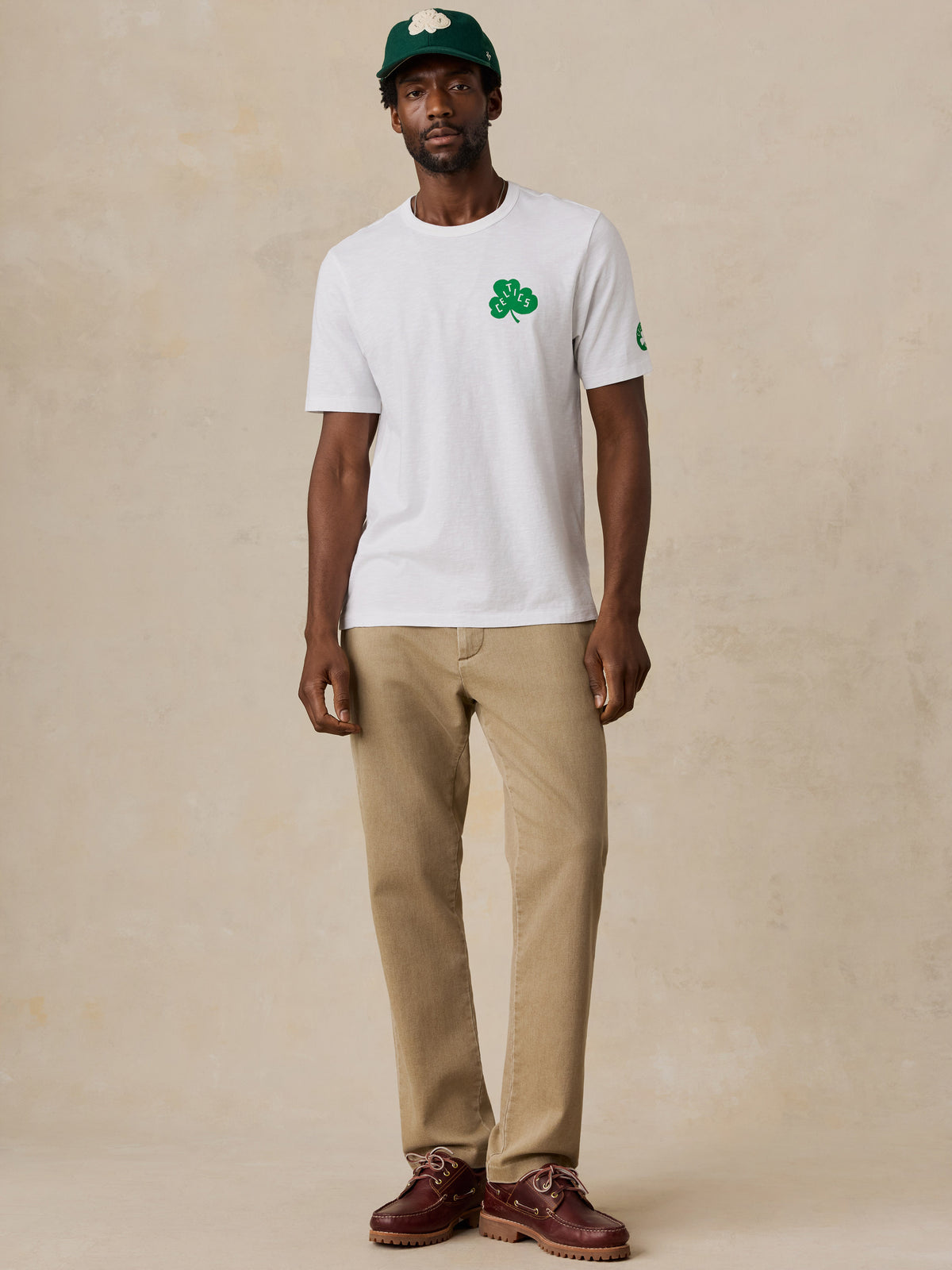 A man wearing a white Boston Celtics tee and chino pants, image 5 of 6
