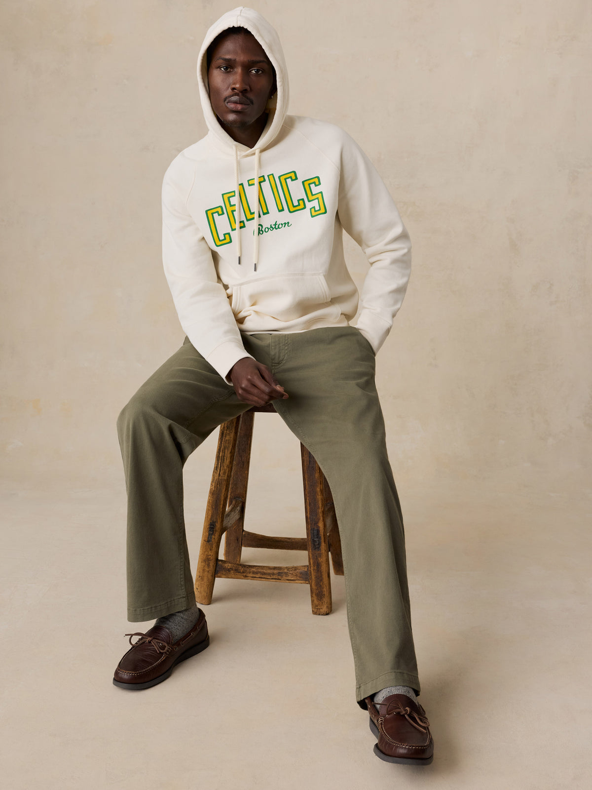A man wearing a white Boston Celtics hoodie and green chino pants, image 6 of 6