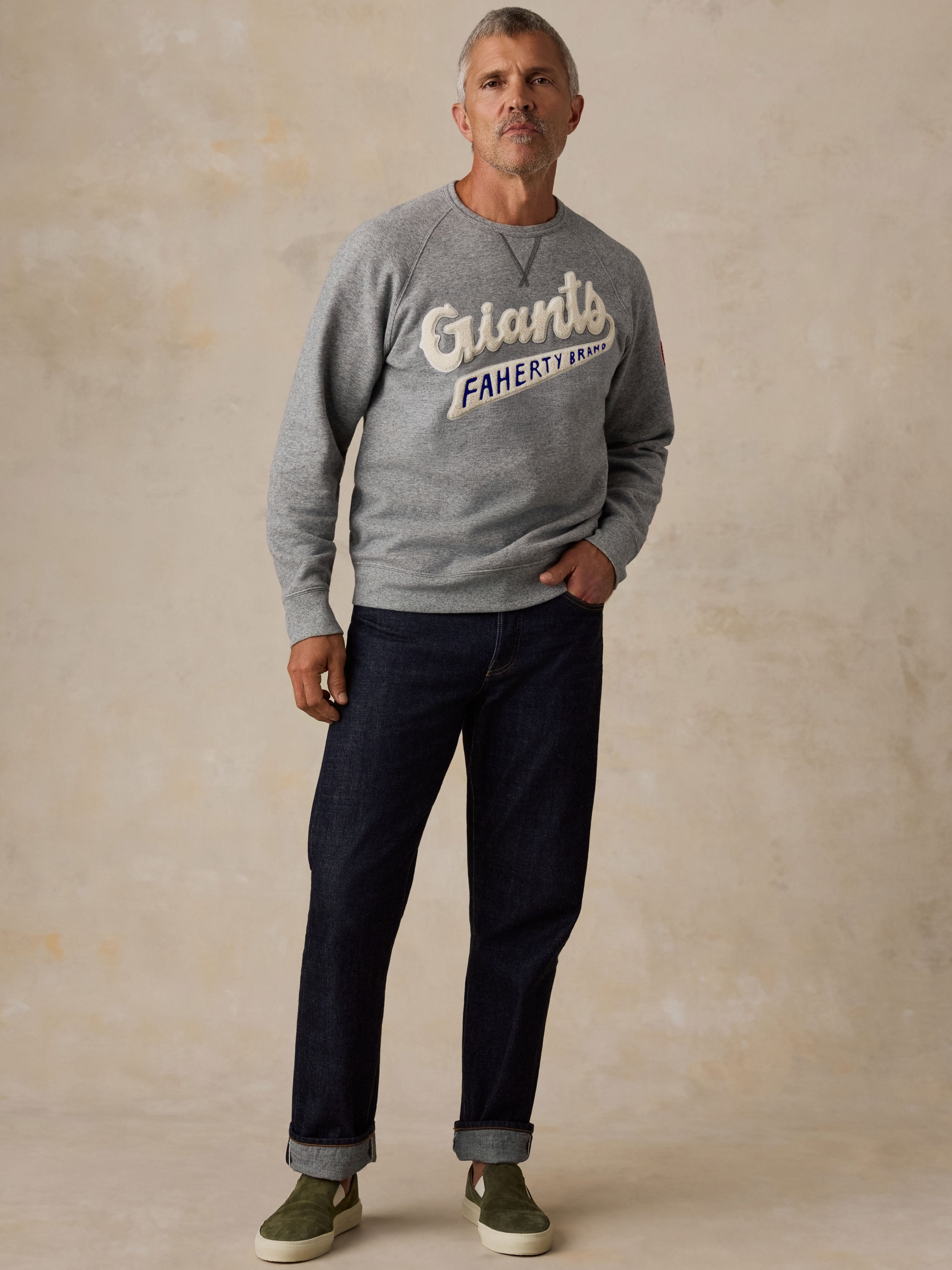 A man wearing a grey NY Giants crewneck, blue jeans, and green sneakers