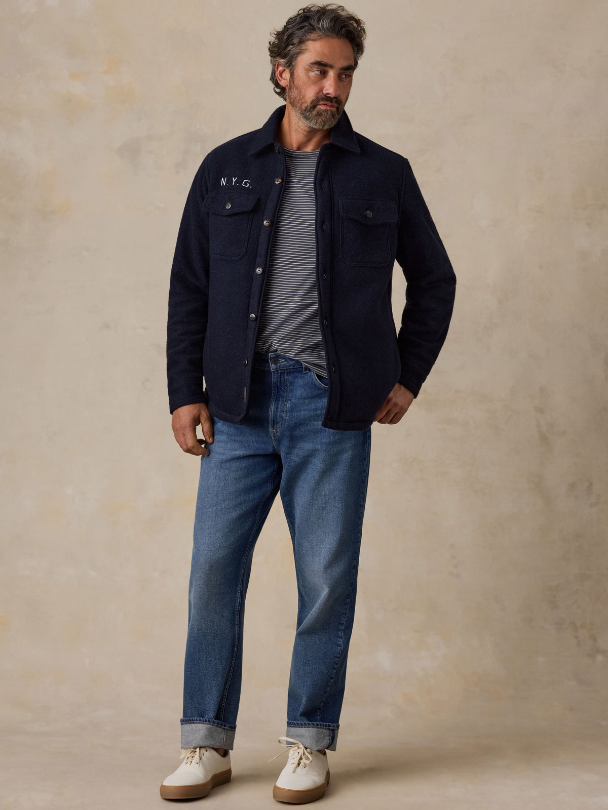 A man wearing a blue NY Giants jacket over a striped tee and blue jeans with white sneakers, image 3 of 5